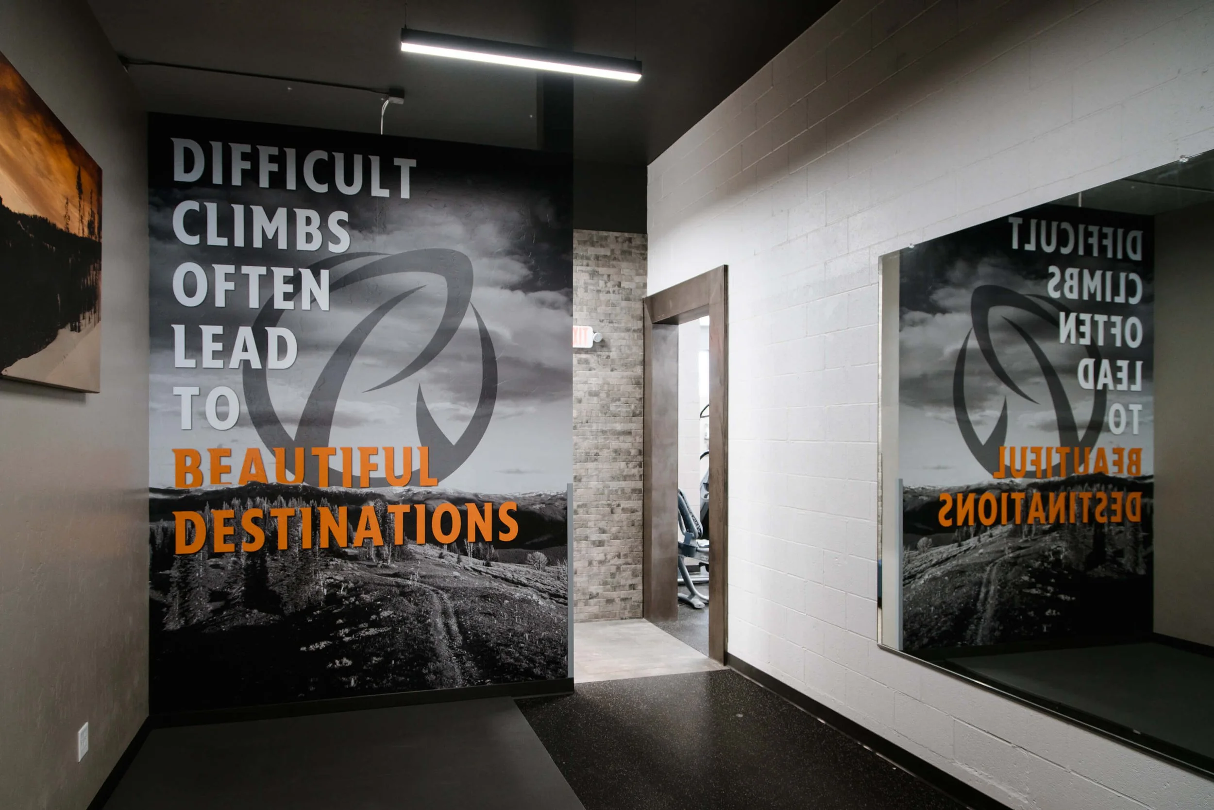 Wall wrap with grayscale mountain landscape background and bold orange message: “DIFFICULT CLIMBS OFTEN LEAD TO BEAUTIFUL DESTINATIONS” in a gym corridor. Installed by The Wrap Agency at the Ridge Athletic Club in Bozeman, Montana.