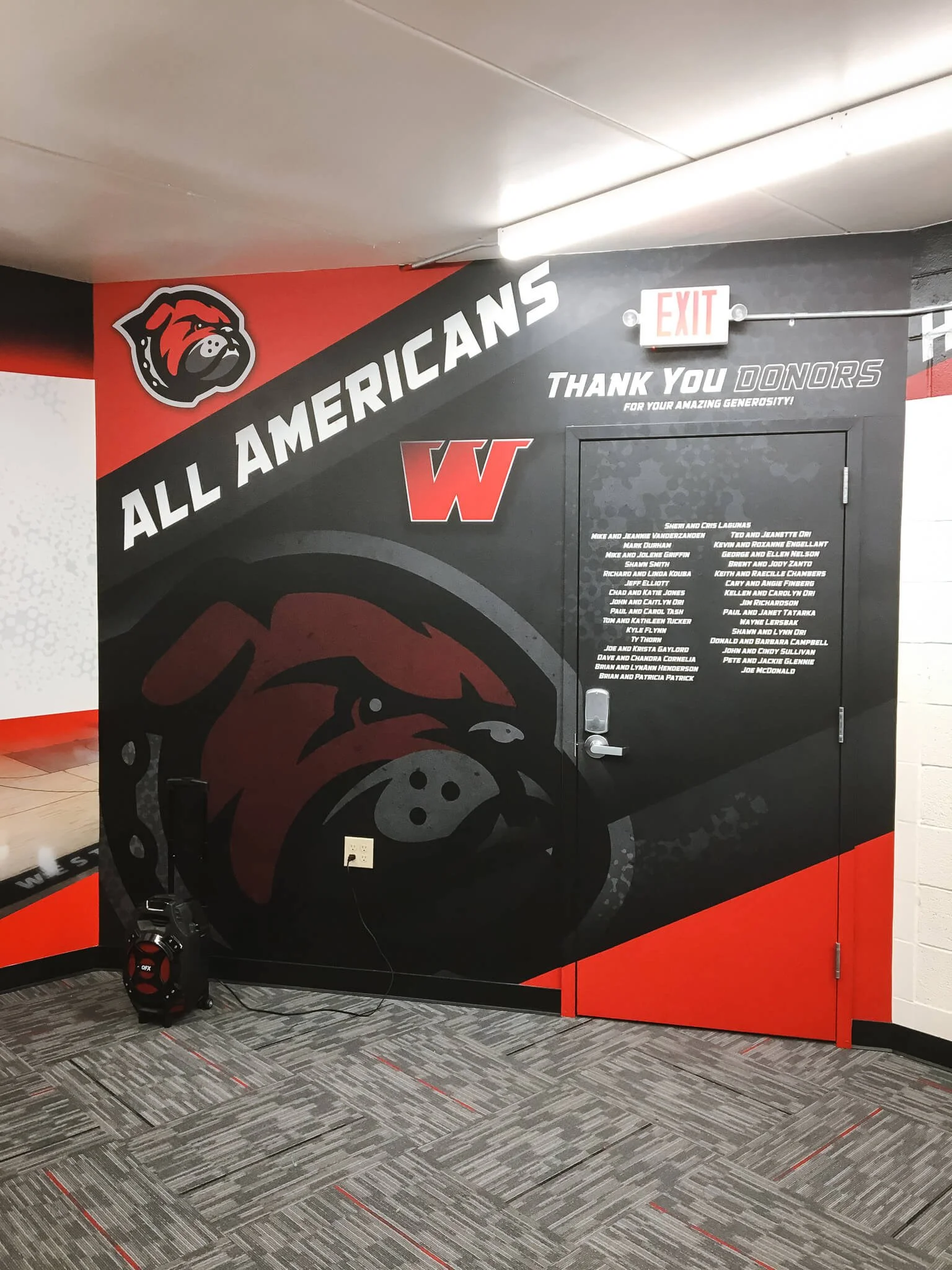 Bold, sports-inspired wall wrap in a school athletic facility, featuring a bulldog mascot, “All Americans” lettering, and a donor thank-you list – installed at UM Western in Dillon, MT.