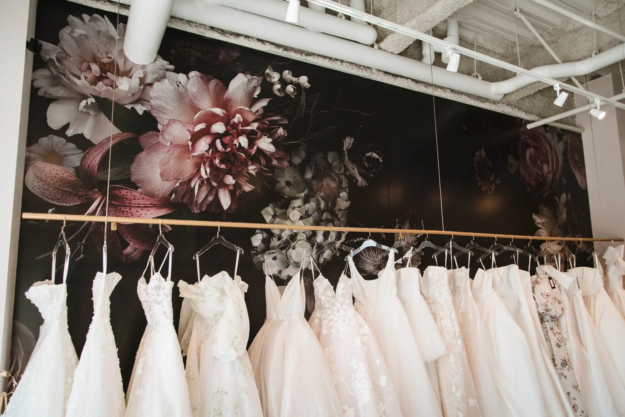 A row of white wedding dresses hanging on a rack in front of a dark moody floral wall wrap. 
