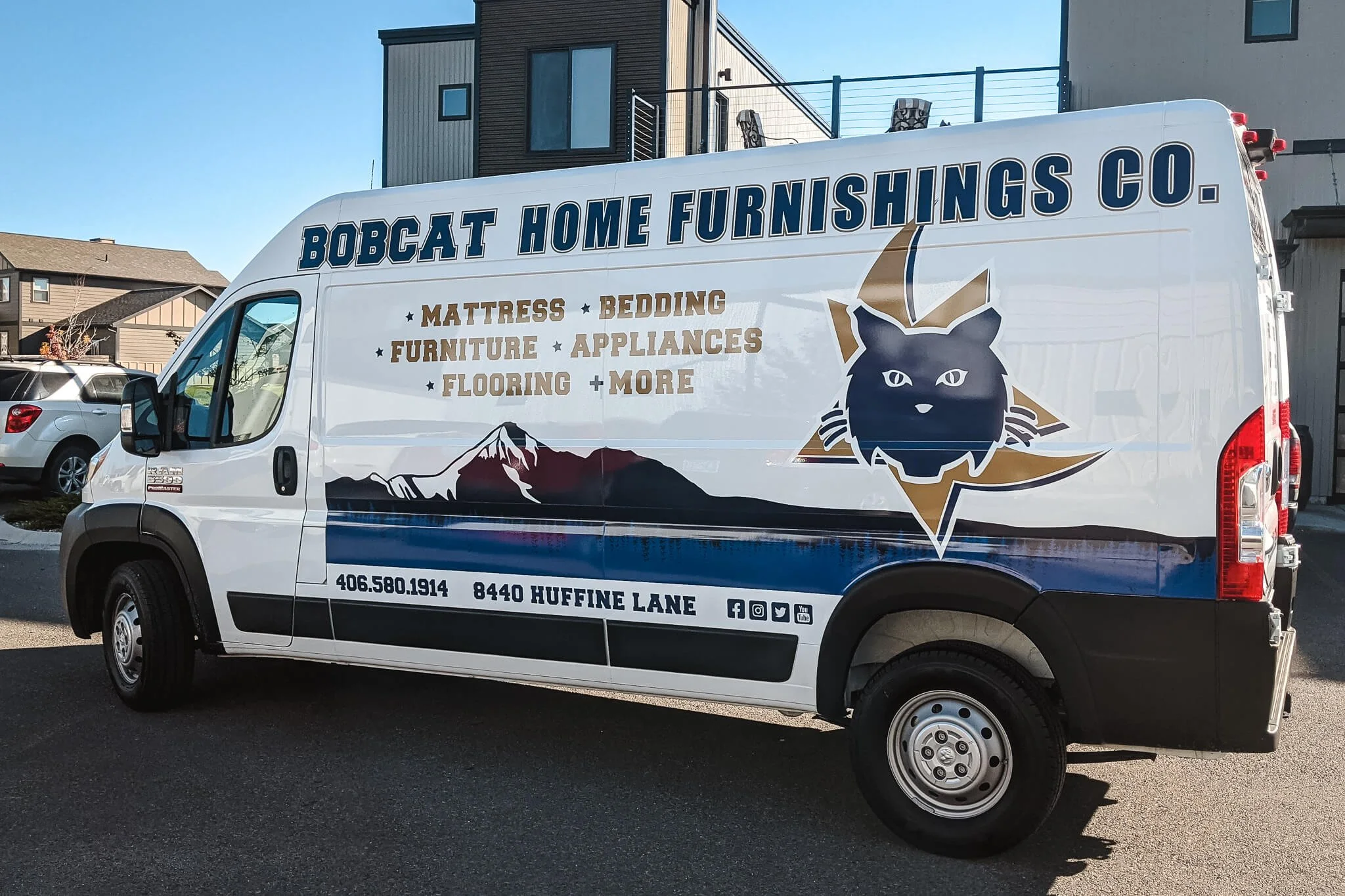 Branded vehicle wrap for Bobcat Home Furnishings Co. on a Ram ProMaster, featuring a blue and gold color scheme with logo, product categories, and mountain graphics. Installed in Bozeman, Montana.