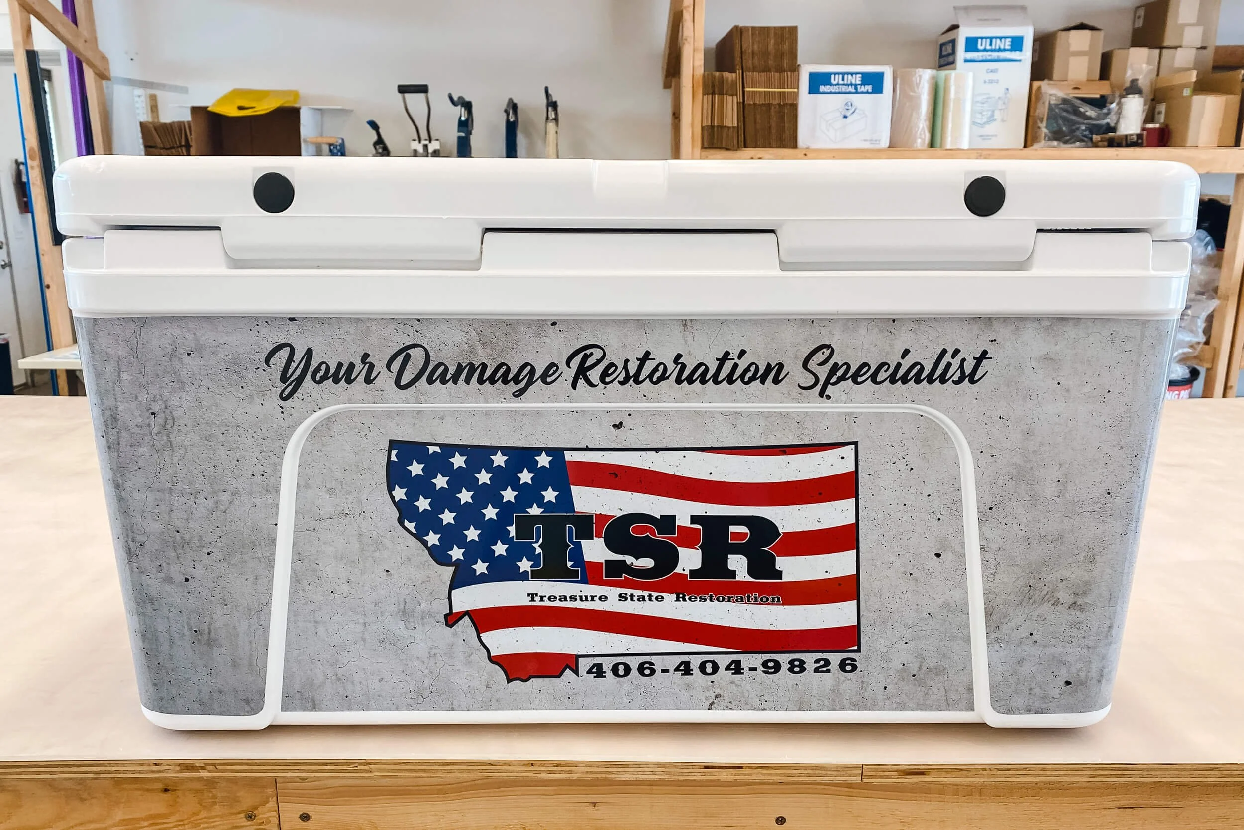 White YETI cooler with concrete texture vinyl wrap with a large American Flag Treasure State Restoration logo and text reading "Your Damage Restoration Specialist"