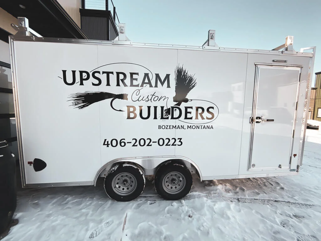Upstream Custom Builders trailer graphics in Bozeman, Montana, featuring black logo and fly fishing design on a white trailer with simple black text showcasing the company’s name and phone number.