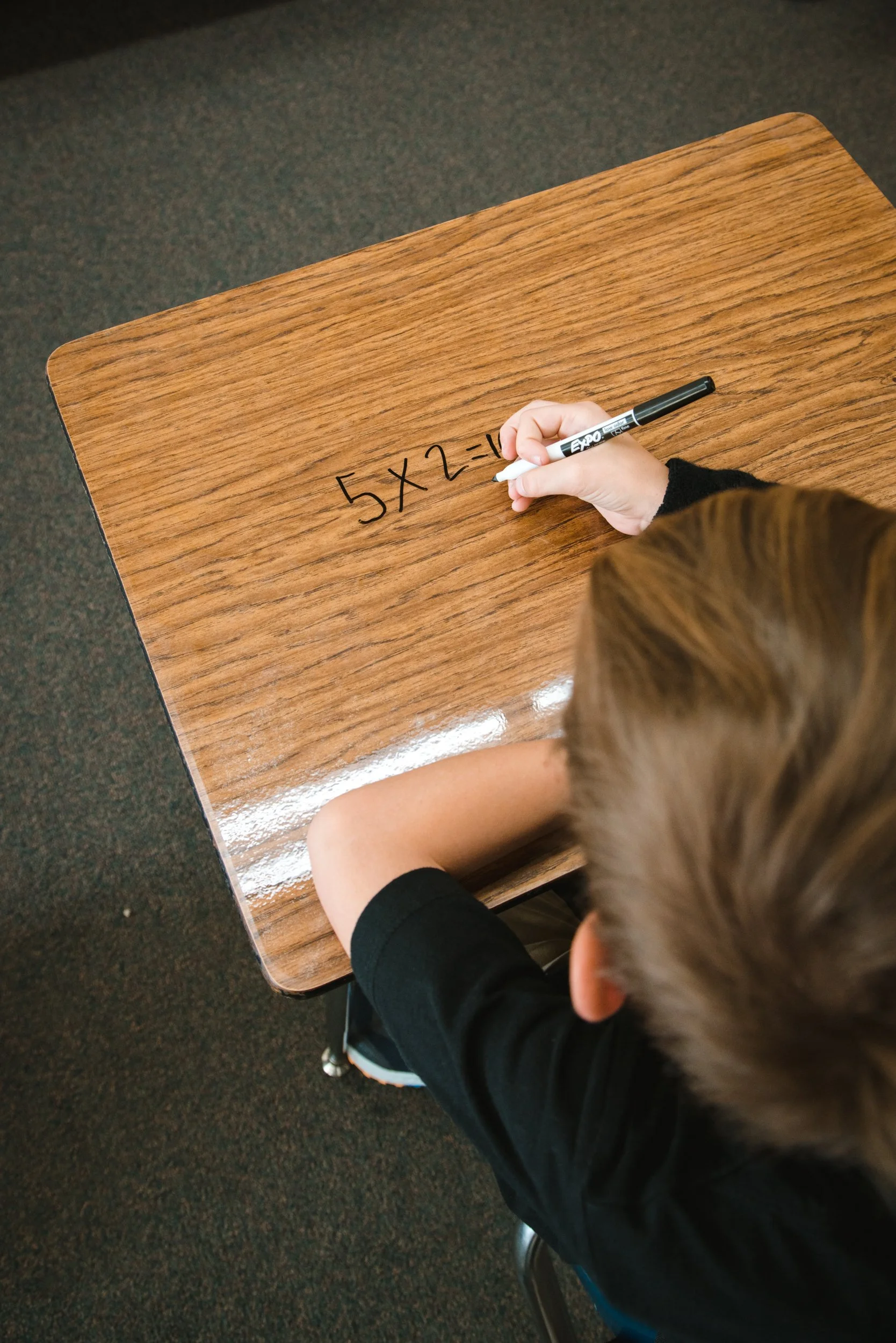 Student writing a multiplication problem on a woodgrain desktop covered with clear dry erase material. 