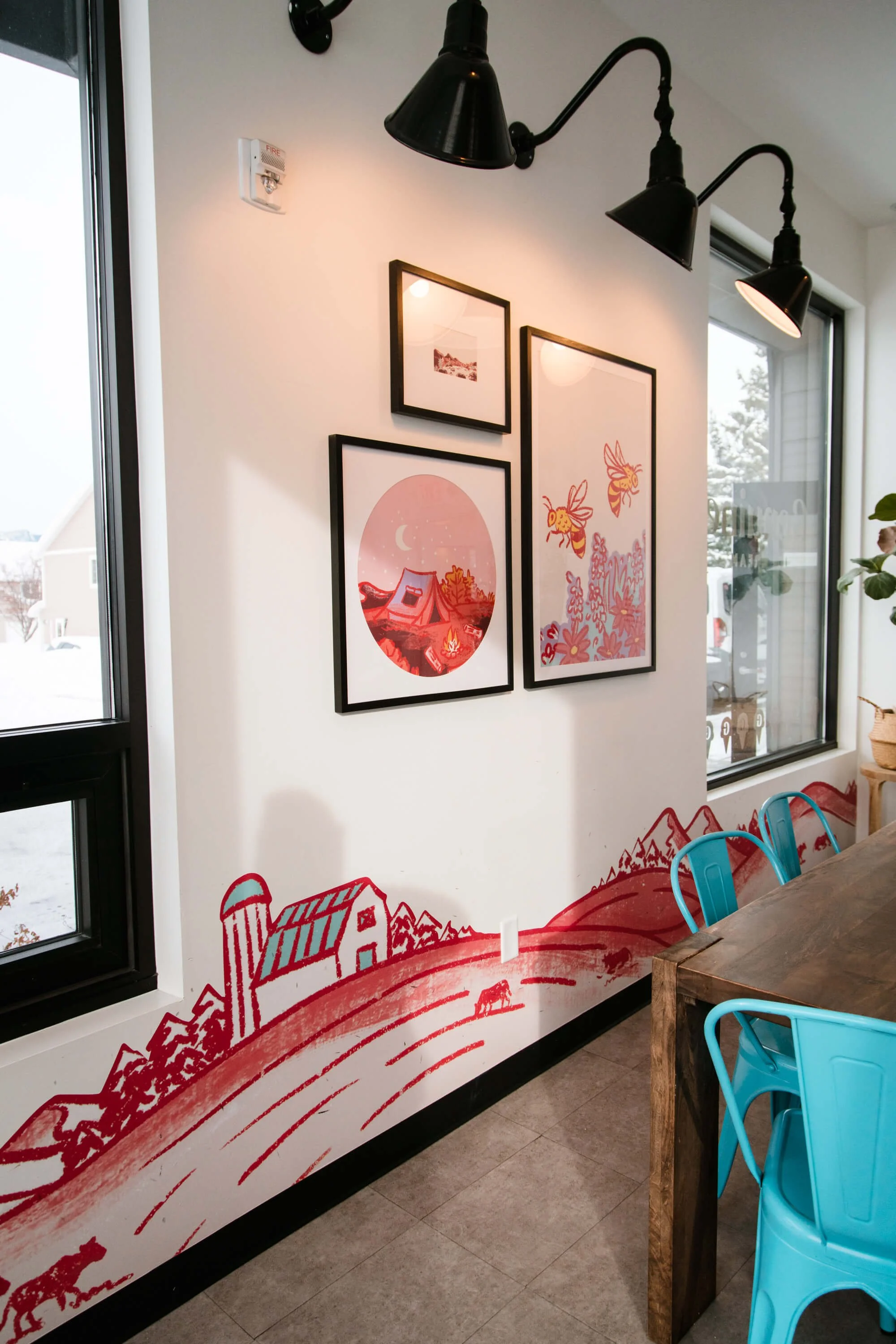 Illustrated red line-art wall wrap of a whimsical rural scene with barns and hills, paired with framed prints and turquoise chairs in a cozy café or restaurant interior. Location: Genuine Ice Cream, Bozeman, MT