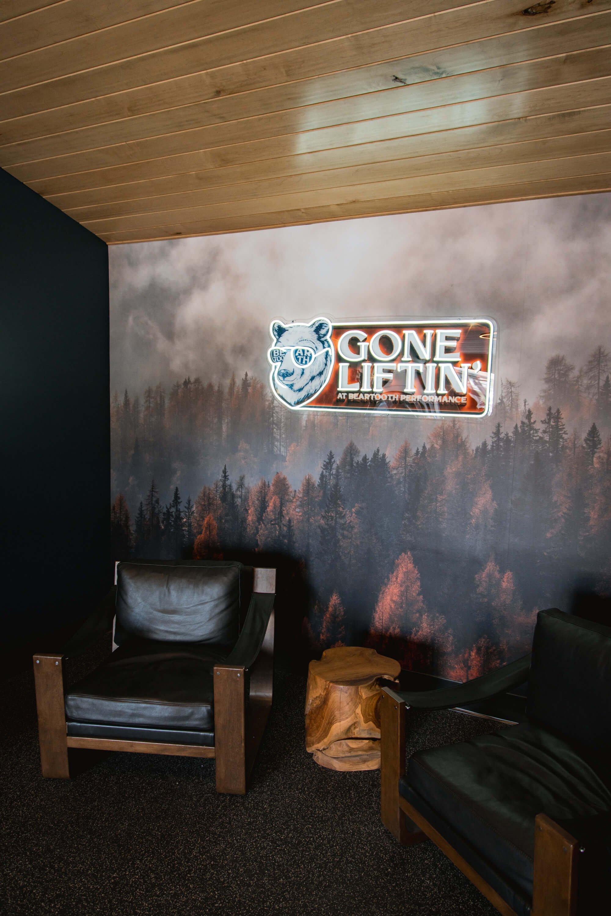 Wall wrap featuring a foggy pine forest backdrop with a neon sign that reads “Gone Liftin’ at Beartooth Performance,” set in a wood-paneled lounge with black leather chairs – installed in Billings, MT.