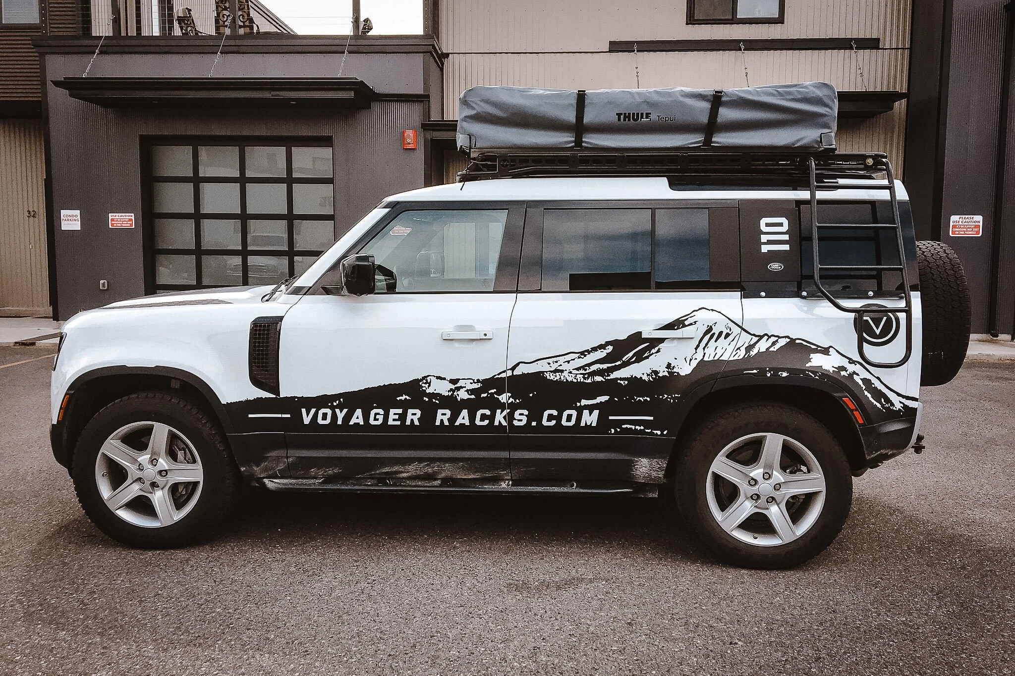 Matte-style vehicle wrap on a white Land Rover Defender 110, featuring mountain silhouettes and bold Voyager Racks logo in high-contrast black. Installed in Bozeman, Montana.