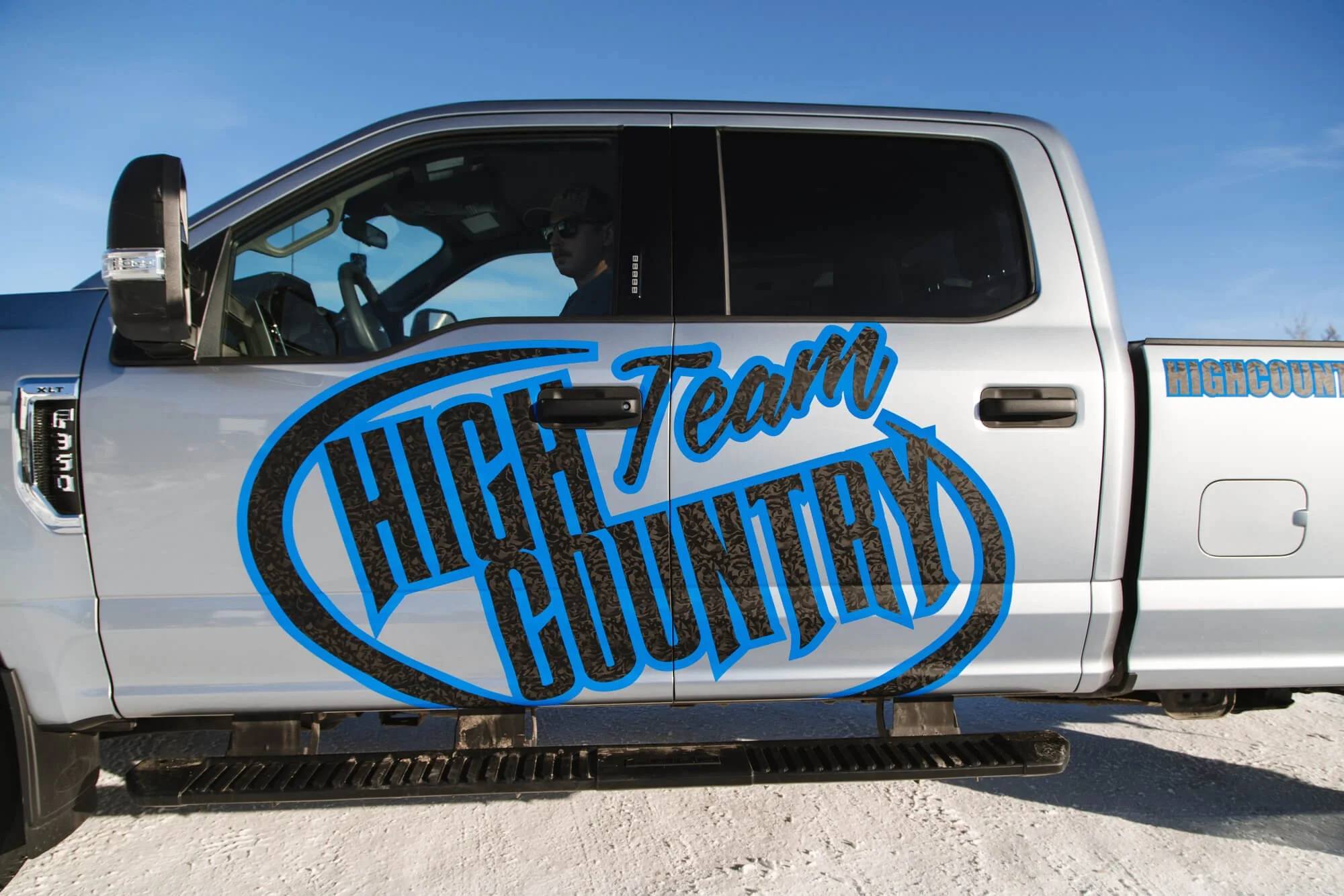 Side view of a silver pickup truck with a blue and black custom vehicle wrap displaying the bold "Team High Country" logo, designed for brand visibility on the road. 