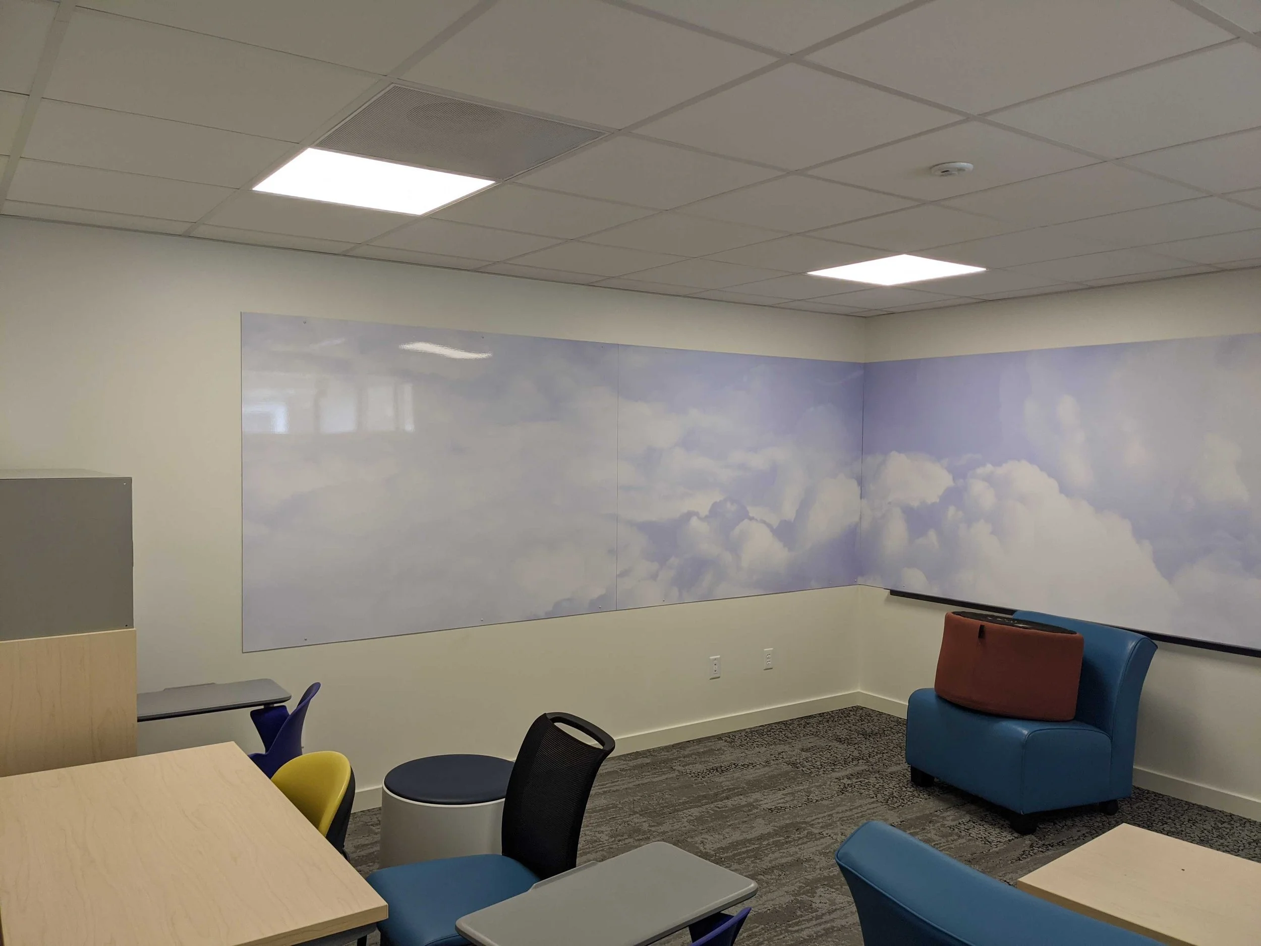 Cloud-patterned dry erase wall graphics wrapping around a corner above blue seating.