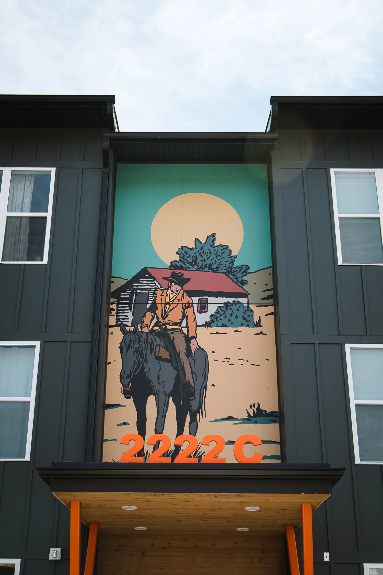 Colorful exterior wall wrap above a townhouse entrance featuring a cowboy on horseback near a cabin under a golden sun, labeled “2222C” in Bozeman, MT.