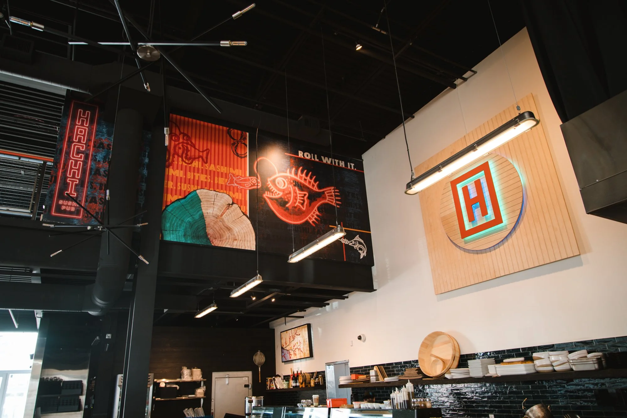 Interior of a Hachi sushi restaurant with vibrant signage, including a custom wall wrap with neon anglerfish, funky graphics and neon Hachi sign.