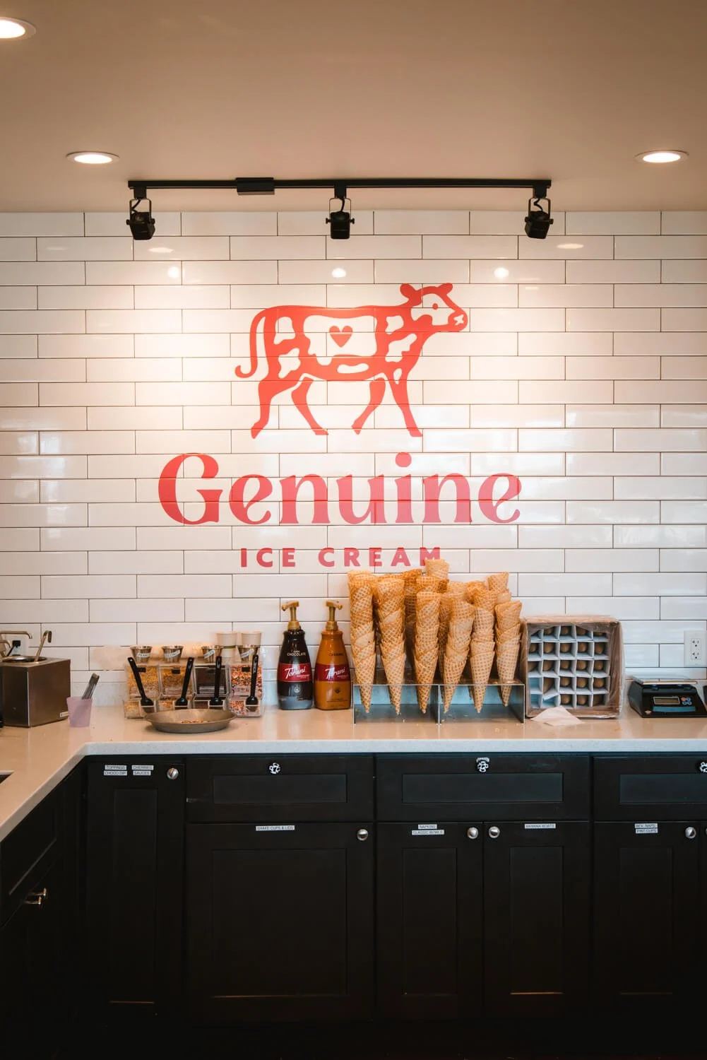 Interior restaurant wall featuring a red “Genuine Ice Cream” logo wall wrap with a cow illustration on white subway tile above a counter lined with cones and toppings in Bozeman, MT.