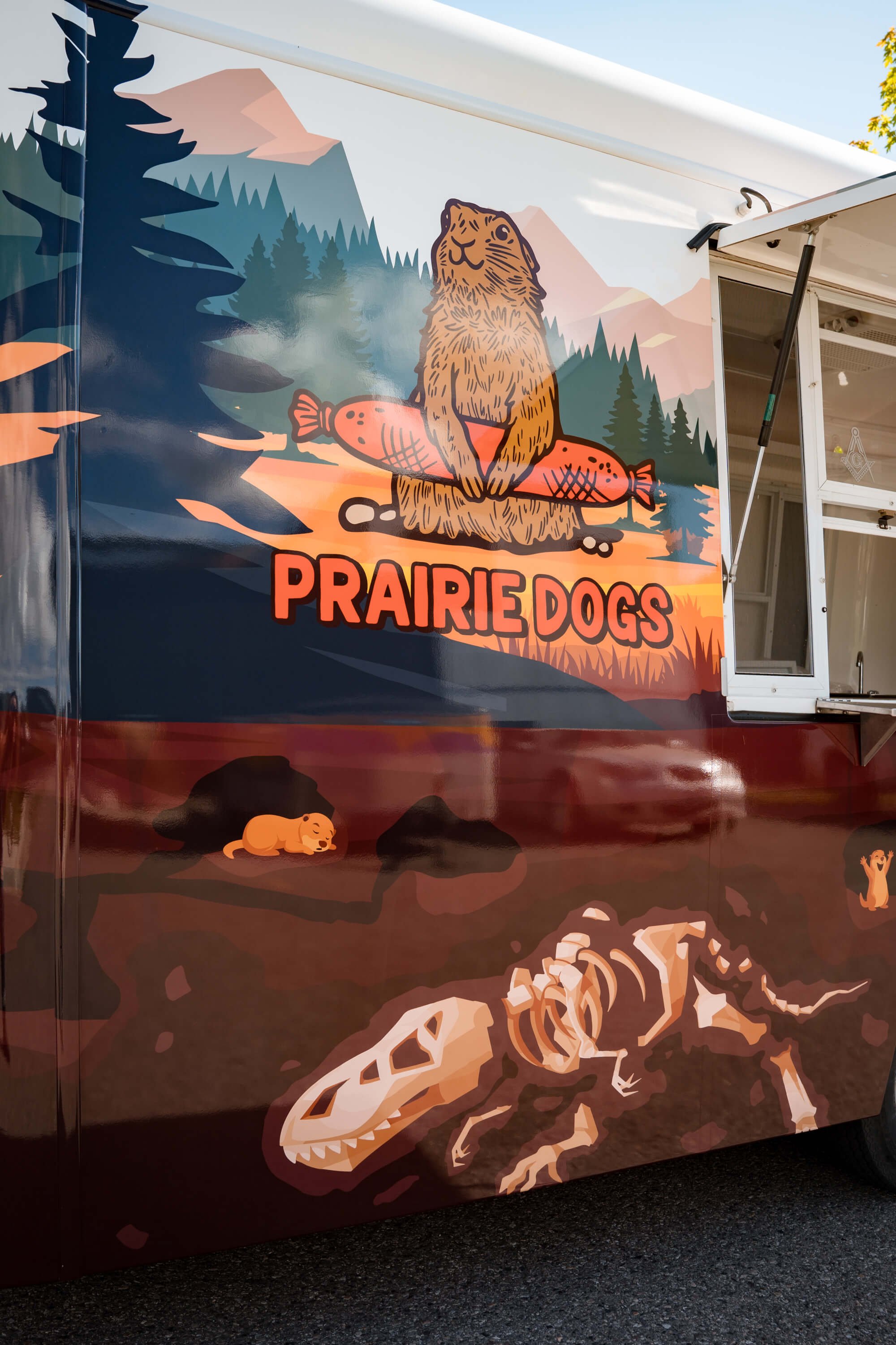 Close-up of custom trailer graphics for Prairie Dogs food trailer in Bozeman, Montana, showing a prairie dog mascot holding sausages, with illustrated forest elements and fossil artwork.