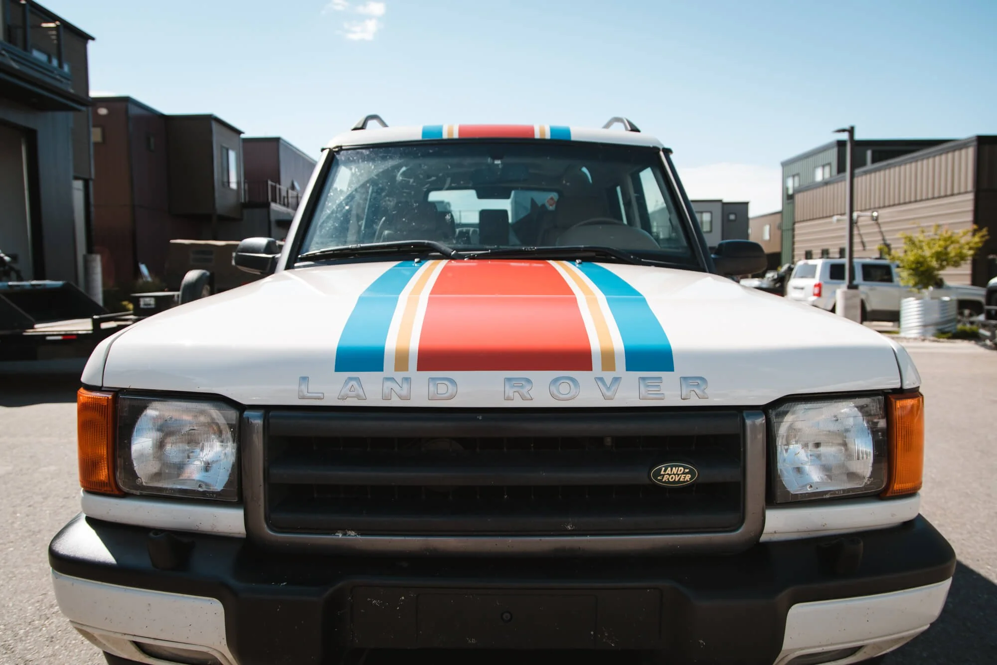 Front view of a white Land Rover with retro-style racing stripes in red, orange, and blue as a partial hood and roof wrap, giving the vehicle a vintage motorsport-inspired aesthetic. Location: Bozeman, MT.
