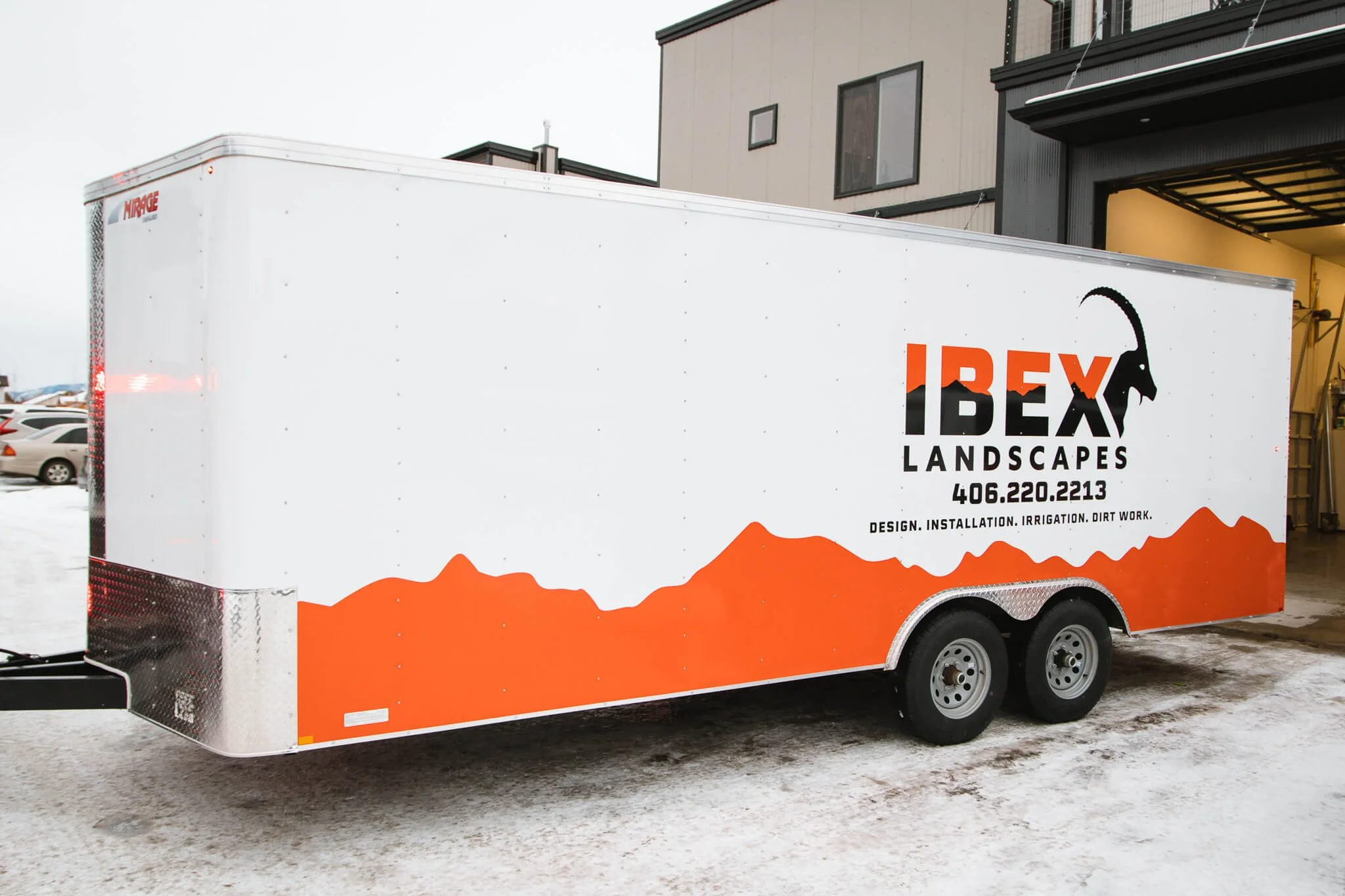 Side view of IBEX Landscapes trailer graphics in Bozeman, Montana, showcasing a clean white wrap with bold orange terrain design and high-contrast branding on a dual-axle enclosed trailer.