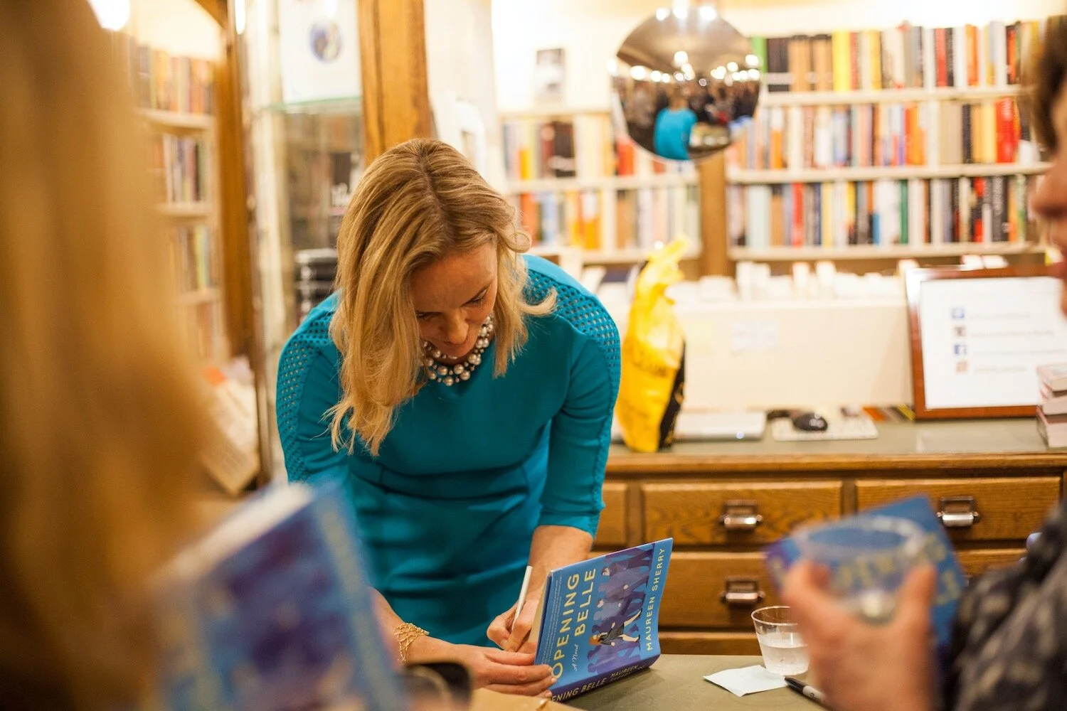 A woman in a teal dress with a pearl necklace signing a book at a bookstore or library event, with bookshelves and a mirror in the background.