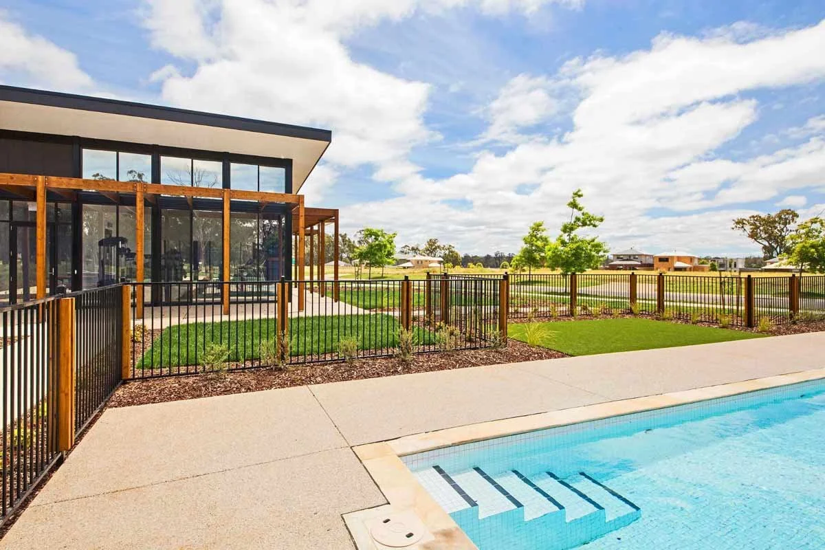 View of backyard with swimming pool, black metal fence, concrete patio, modern house with large glass windows, and a grassy yard with small trees under partly cloudy sky.