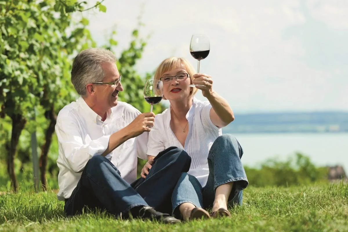 A happy older couple sitting on the grass outdoors, holding glasses of red wine, with a lake and green trees in the background.
