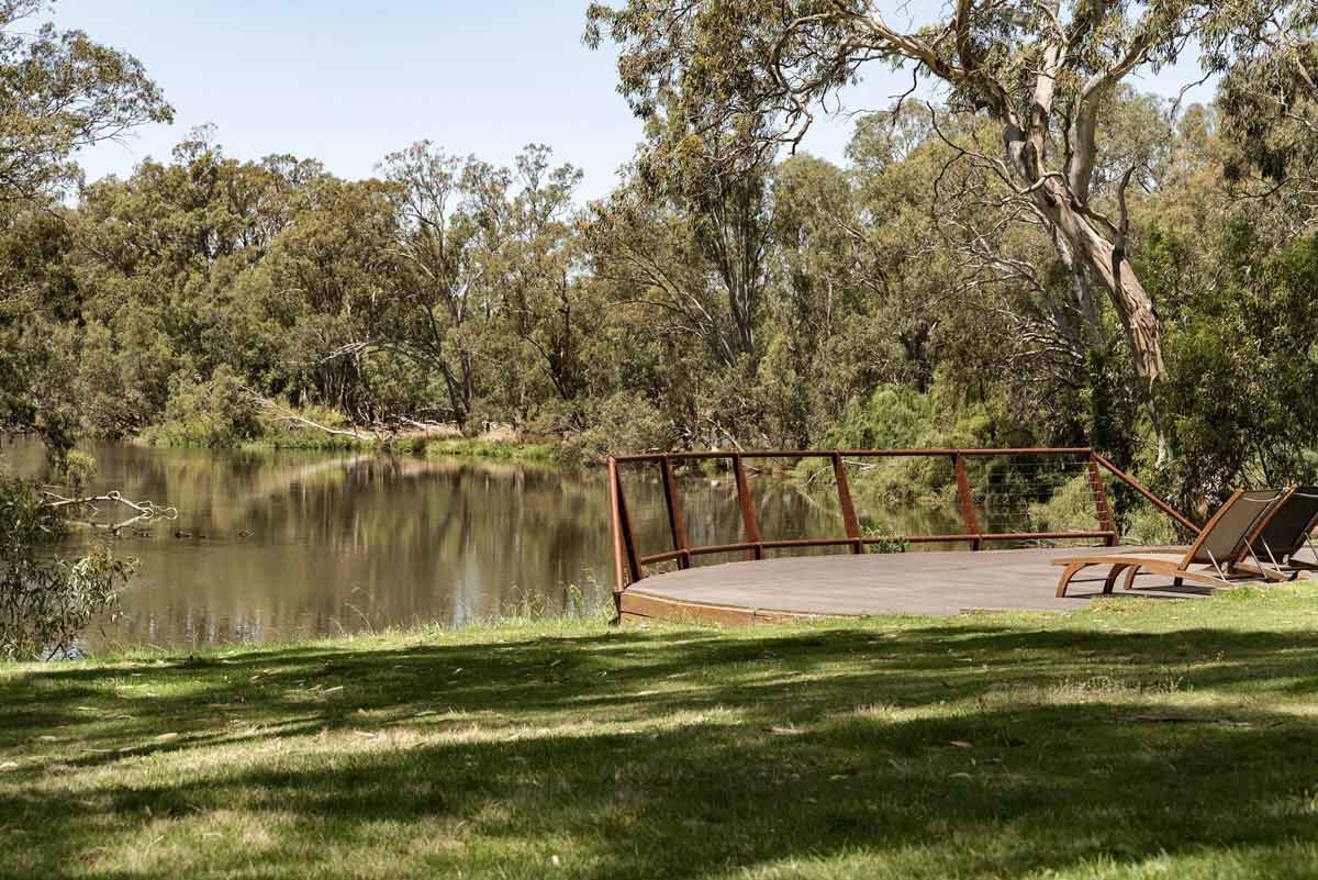 A wooden deck with outdoor lounge chairs by a river, surrounded by green trees under a clear sky.