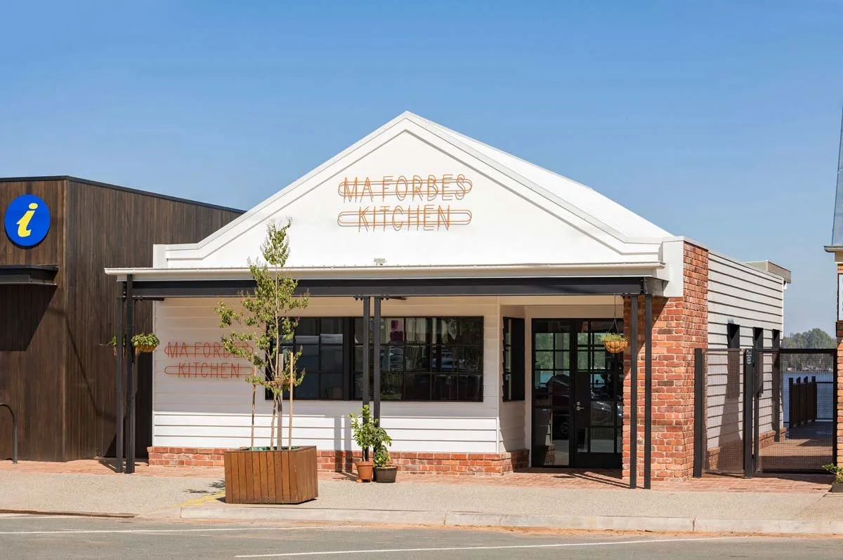 Exterior view of a small restaurant called 'Ma Forbes Kitchen' with white siding, a brick foundation, and a neon sign on the front. A tree in a planter and smaller potted plants are outside, with a sidewalk and street in the foreground.