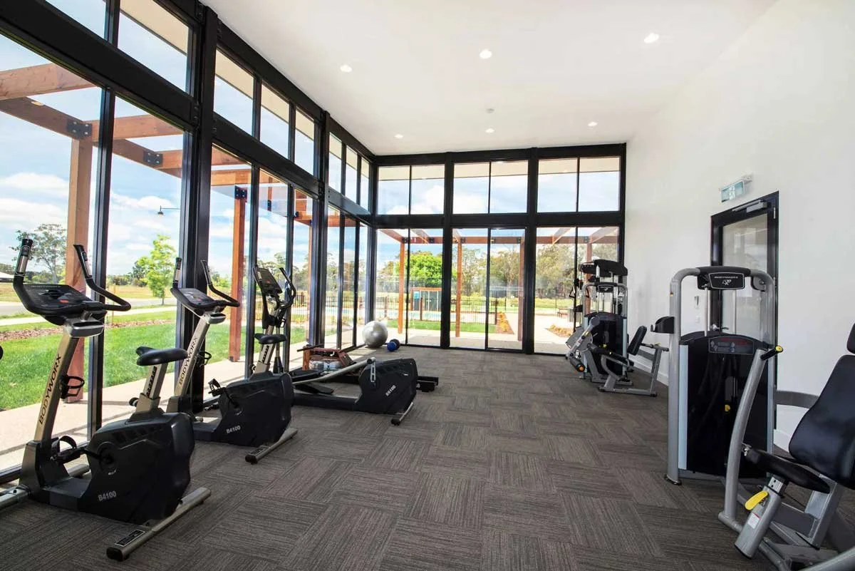 Indoor gym with exercise equipment, including stationary bikes, elliptical trainers, and weight machines, with large glass windows showing an outdoor park and blue sky.