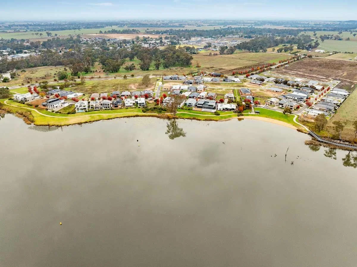 Aerial view of a residential neighborhood with houses, trees, and a small lake in the foreground, set against a rural landscape
