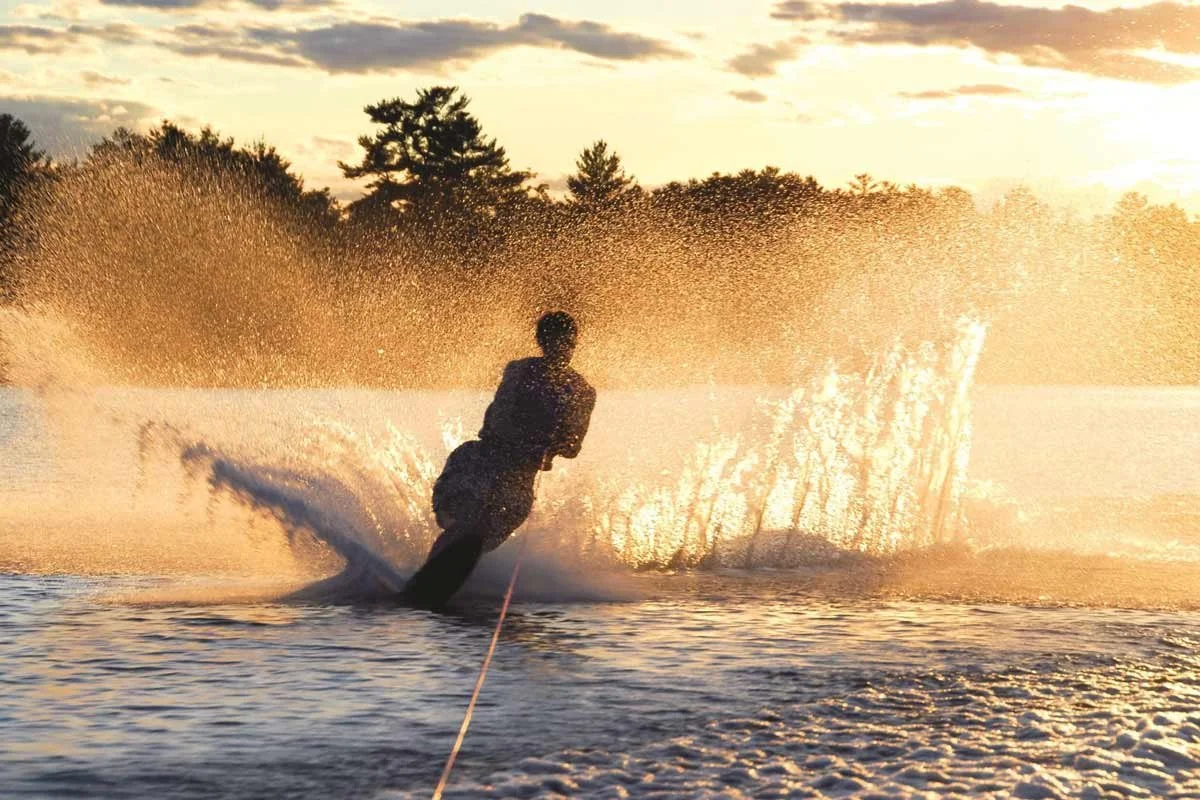 A person waterskiing on a lake during sunset, creating a splash with water spray and silhouetted against the sky with clouds and trees in the background.