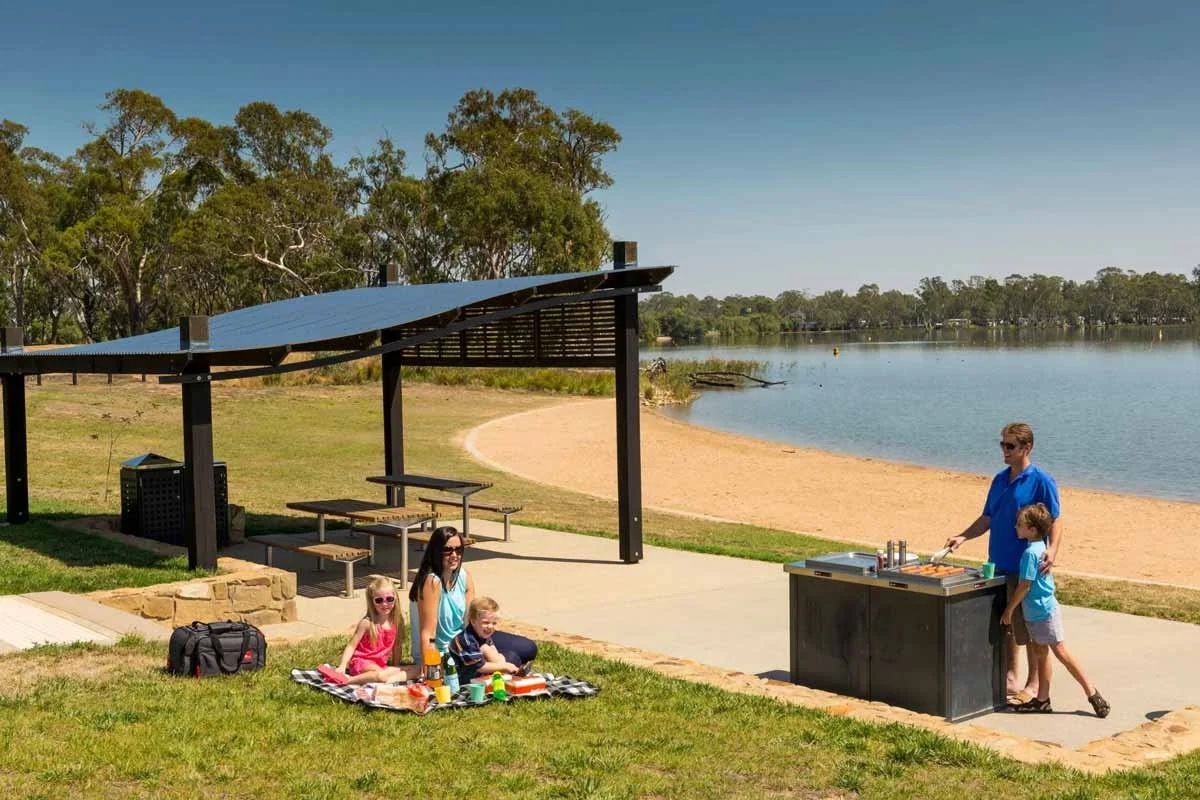 A family enjoying a picnic and barbecue by a lake on a sunny day, with trees in the background and a sandy beach nearby.