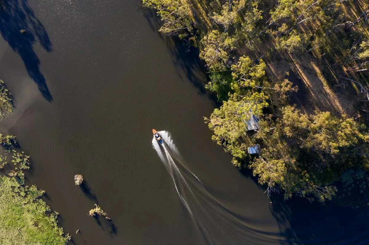 Aerial view of a speedboat on a dark river, bordered by dense green trees and small structures, with shadows cast by trees on the water.