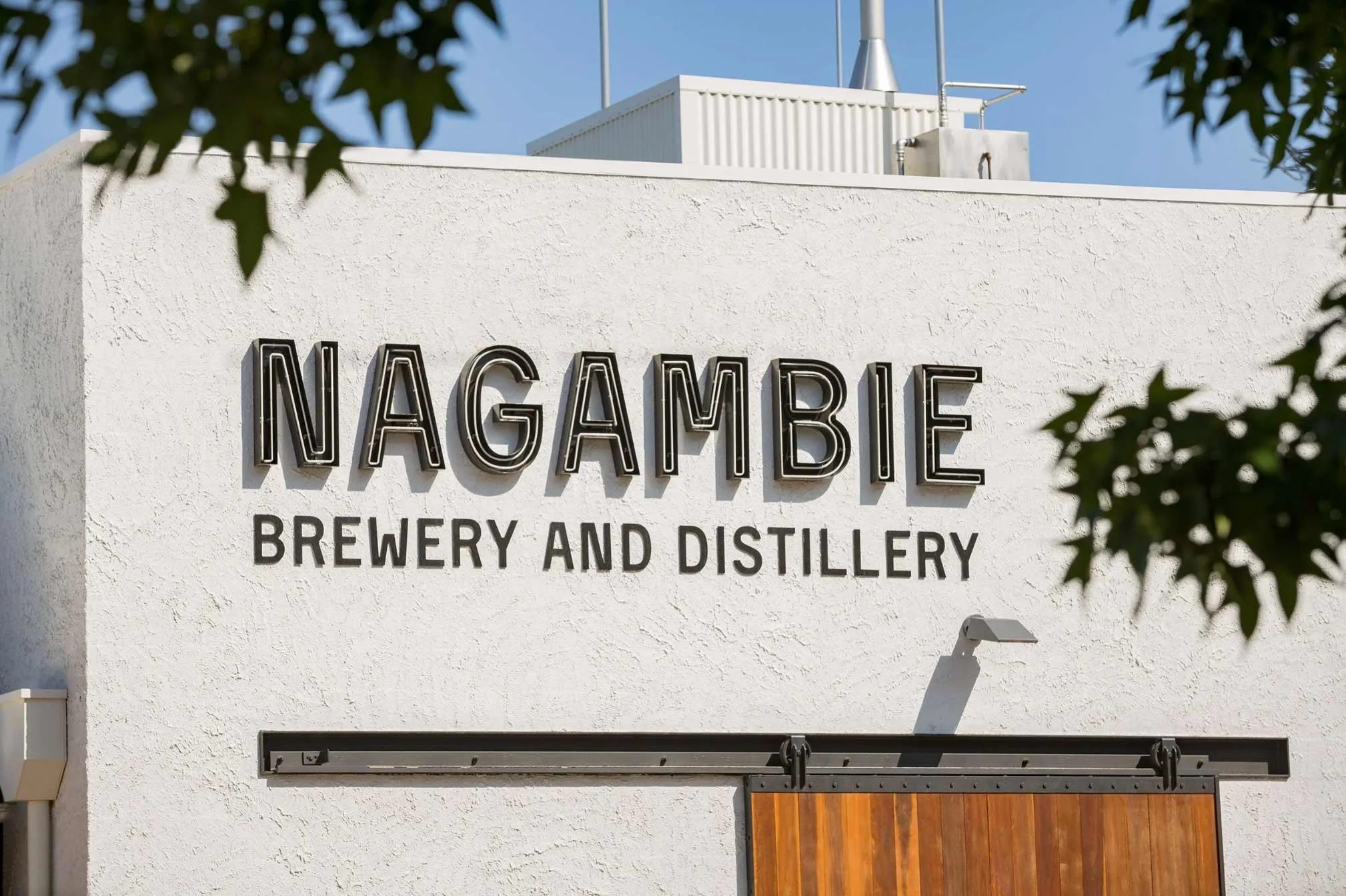 Exterior sign for Nagambie Brewery and Distillery on a white stucco wall with trees partially framing the view.