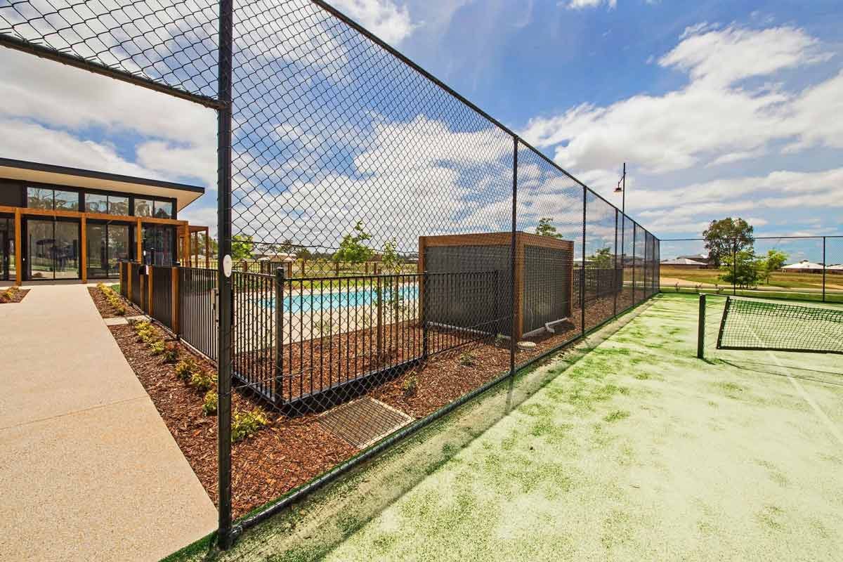 Fenced outdoor area with a swimming pool and a tennis court, part of a modern building complex, under a partly cloudy sky.