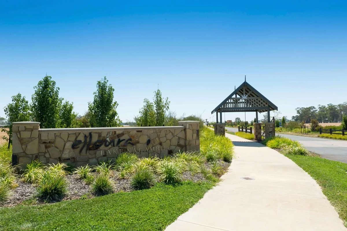 Entrance sign reading "The Harbour Lake N Lake" with a stone wall and landscaped plants, a paved sidewalk leading to a wooden pavilion, and a road in the background under a clear blue sky.