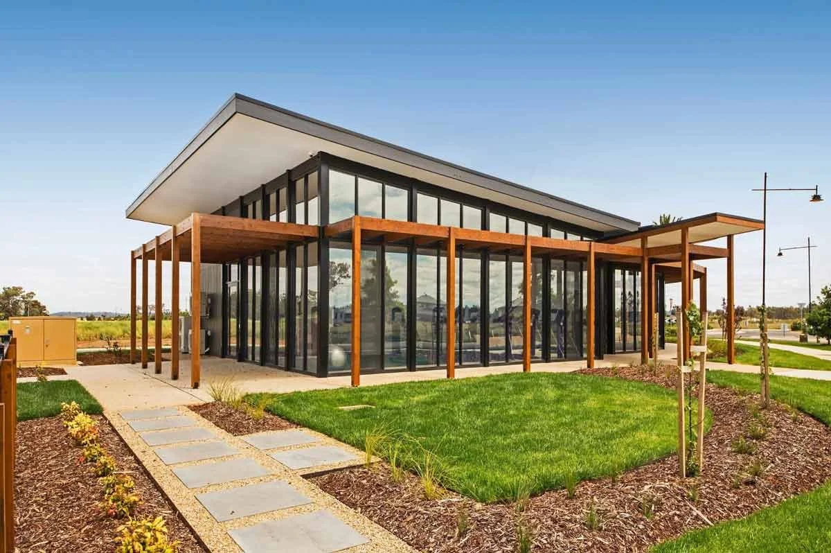 Modern glass and wood building with a flat roof, surrounded by a landscaped yard with grass, plants, and a stone pathway.