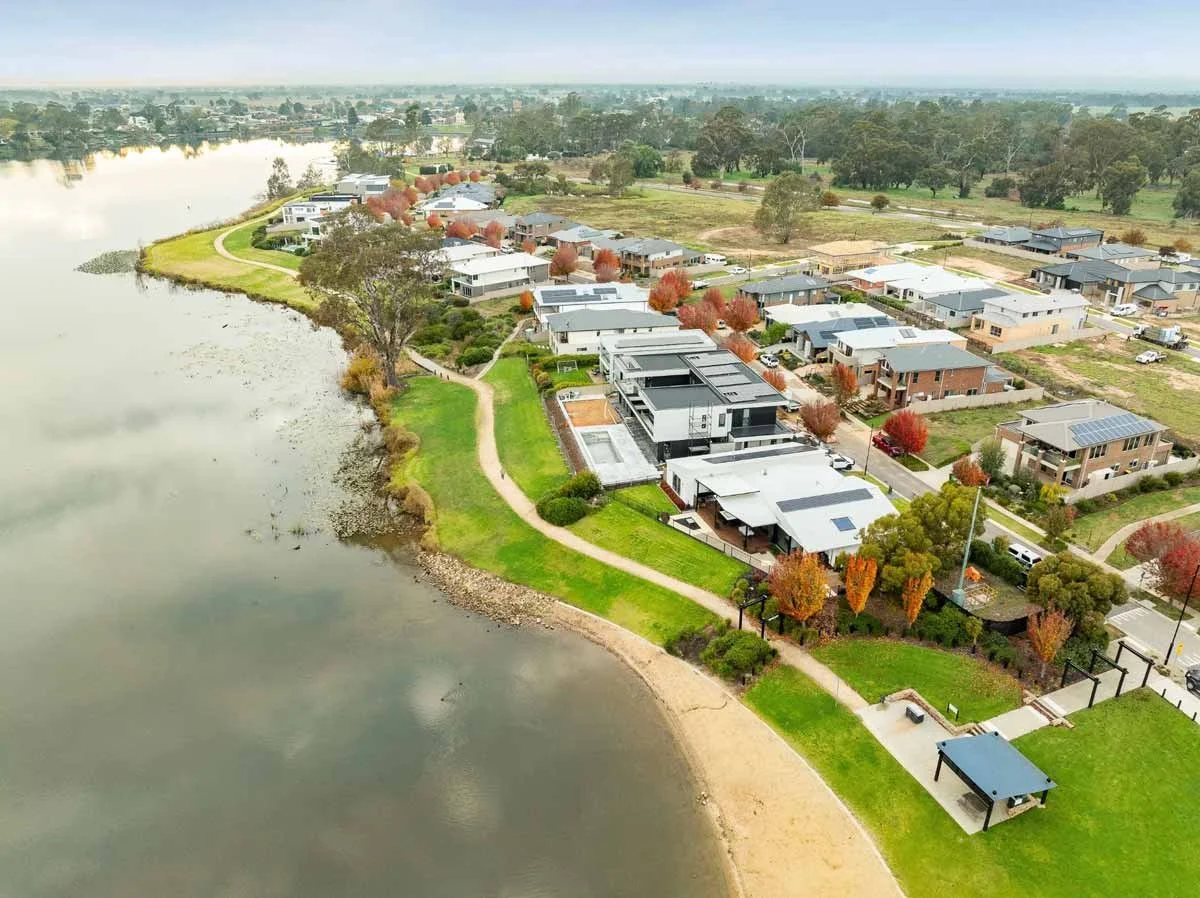 Aerial view of a lakeside residential community with modern houses, green lawns, walking paths, and trees with autumn foliage.