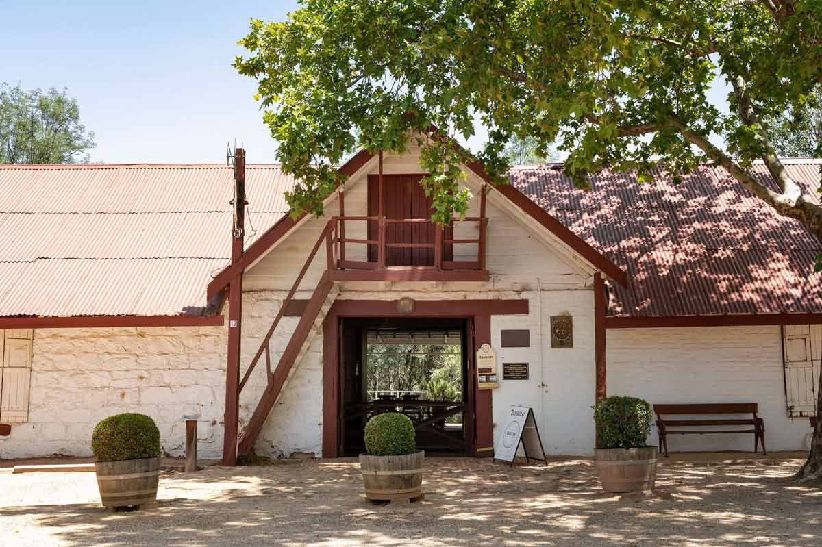 A rustic white building with a red corrugated metal roof, a small second-floor balcony, and a staircase leading to it, surrounded by potted plants and outdoor seating, with a large tree providing shade.