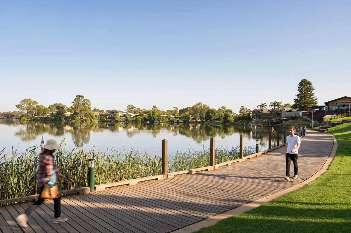 A lakeside scene with a wooden walkway, two people walking along it, and houses across the water under a clear sky.
