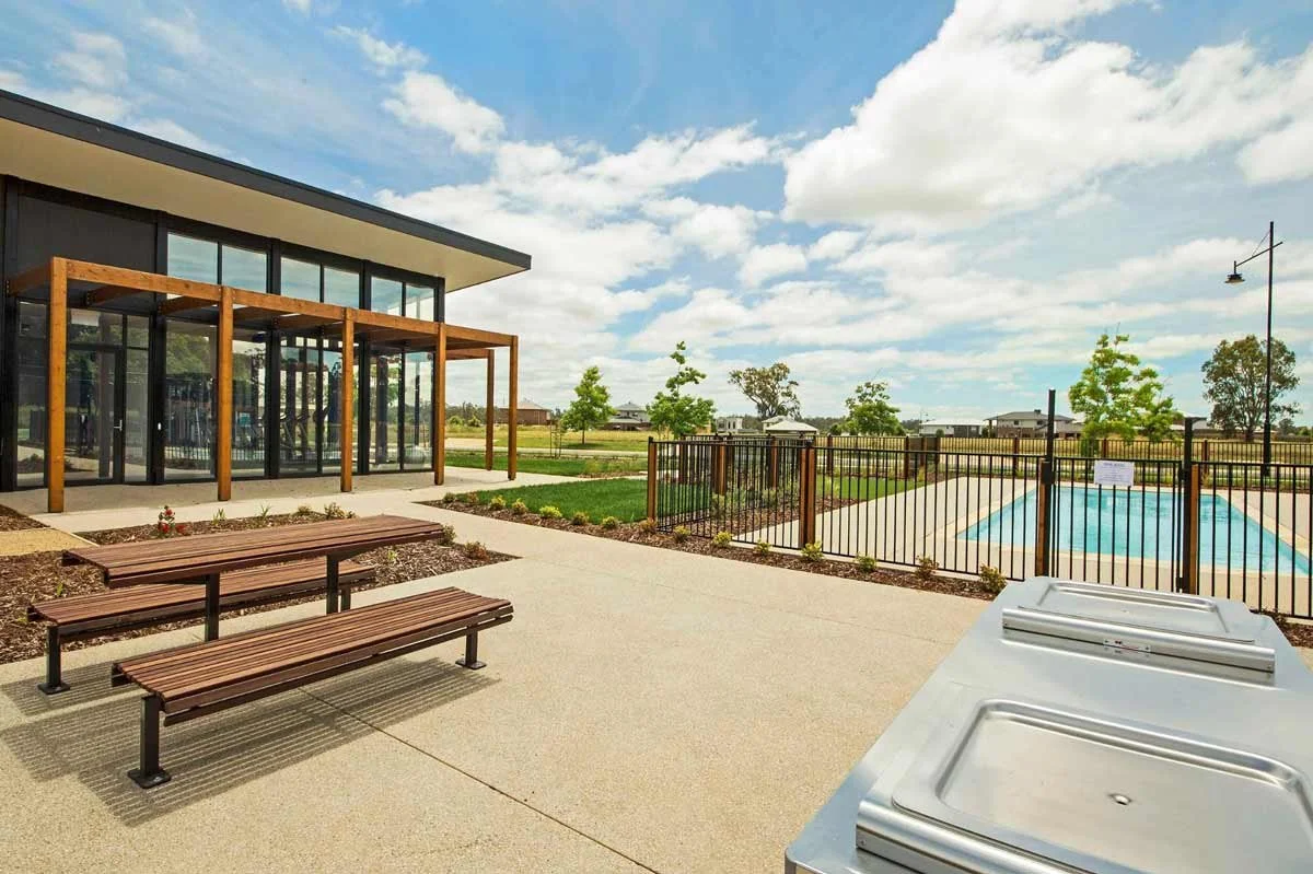 Outdoor community pool area with a fenced-in pool, benches, a modern building with glass walls, and surrounding greenery under a partly cloudy sky.
