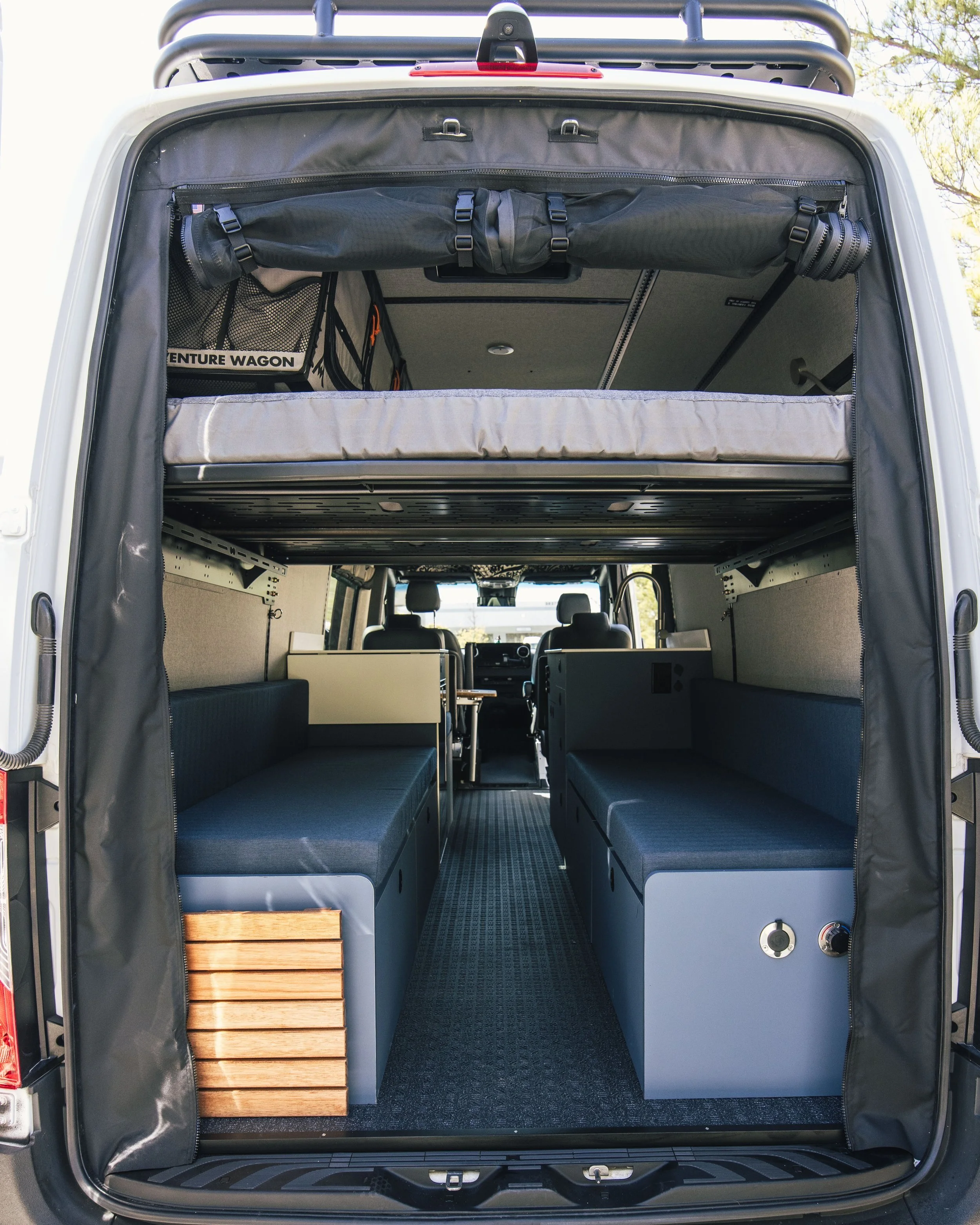 The back of an adventure van with two parallel bench seats, a cargo area underneath, and a loft bed above. The interior is organized for travel and camping.