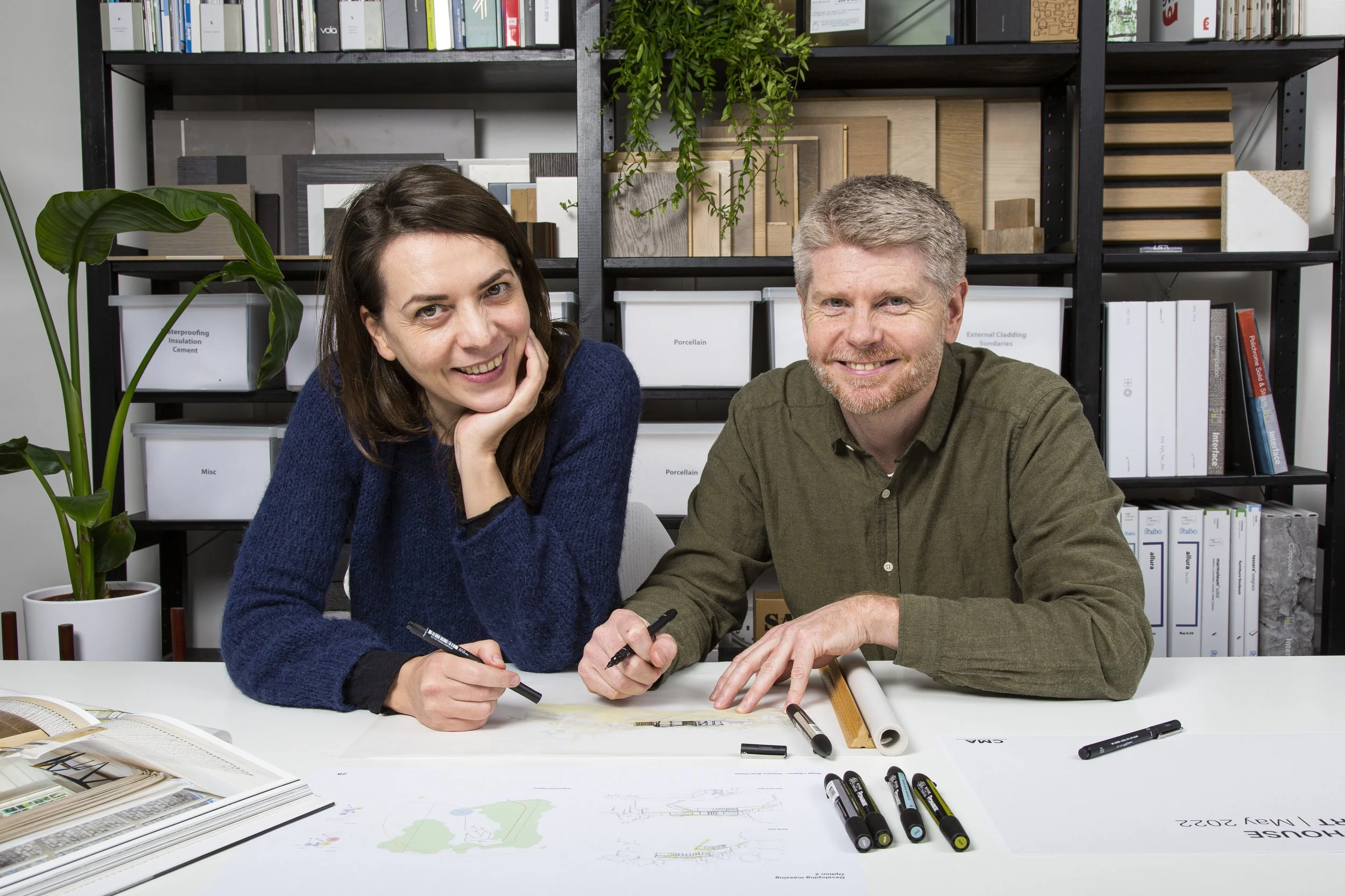 Greg and Daniela, co-founders of Studio CMA, sitting at their design table with drawings and material samples.