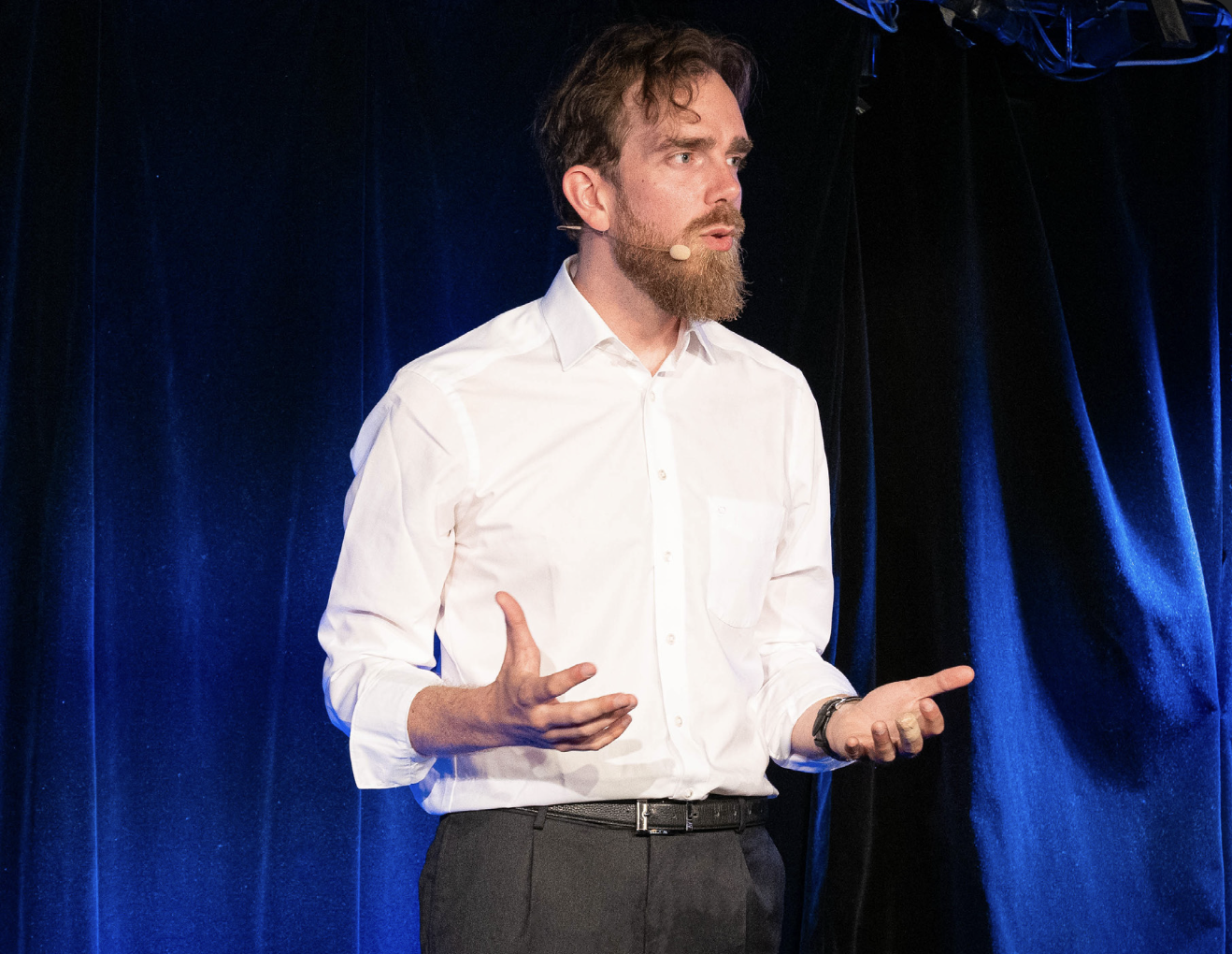 Homme en chemise blanche parlant lors d'une présentation avec un microphone cravate noire et arrière-plan en velours bleu.