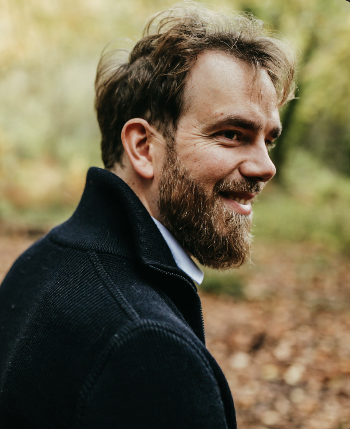 Un homme souriant avec une barbe et des cheveux bruns, portant une veste foncée, se tient dans un environnement forestier en automne.