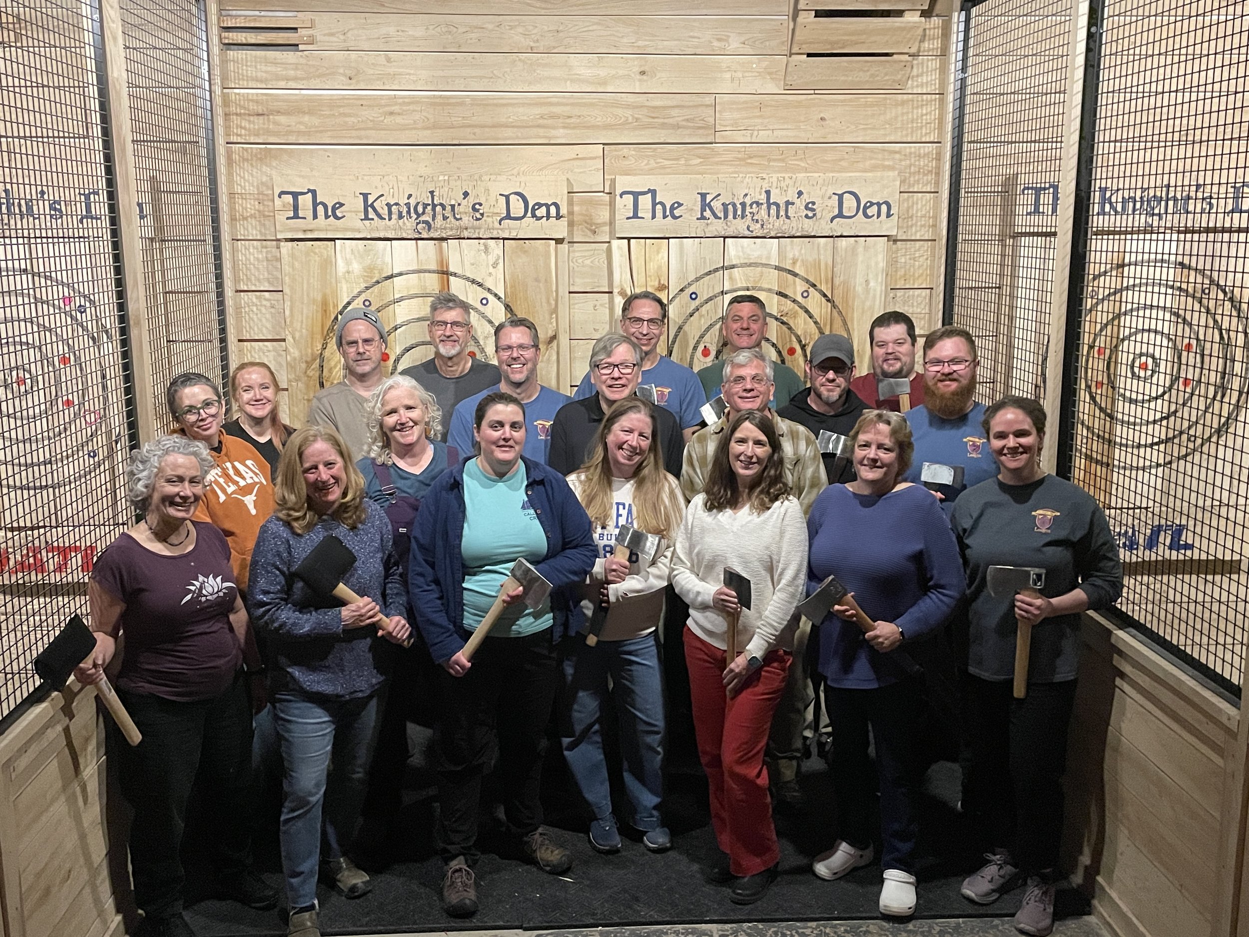 Group of people holding axe-throwing tools in a target hall with wooden decor and netting on the sides, labeled 'The Knight's Den'.