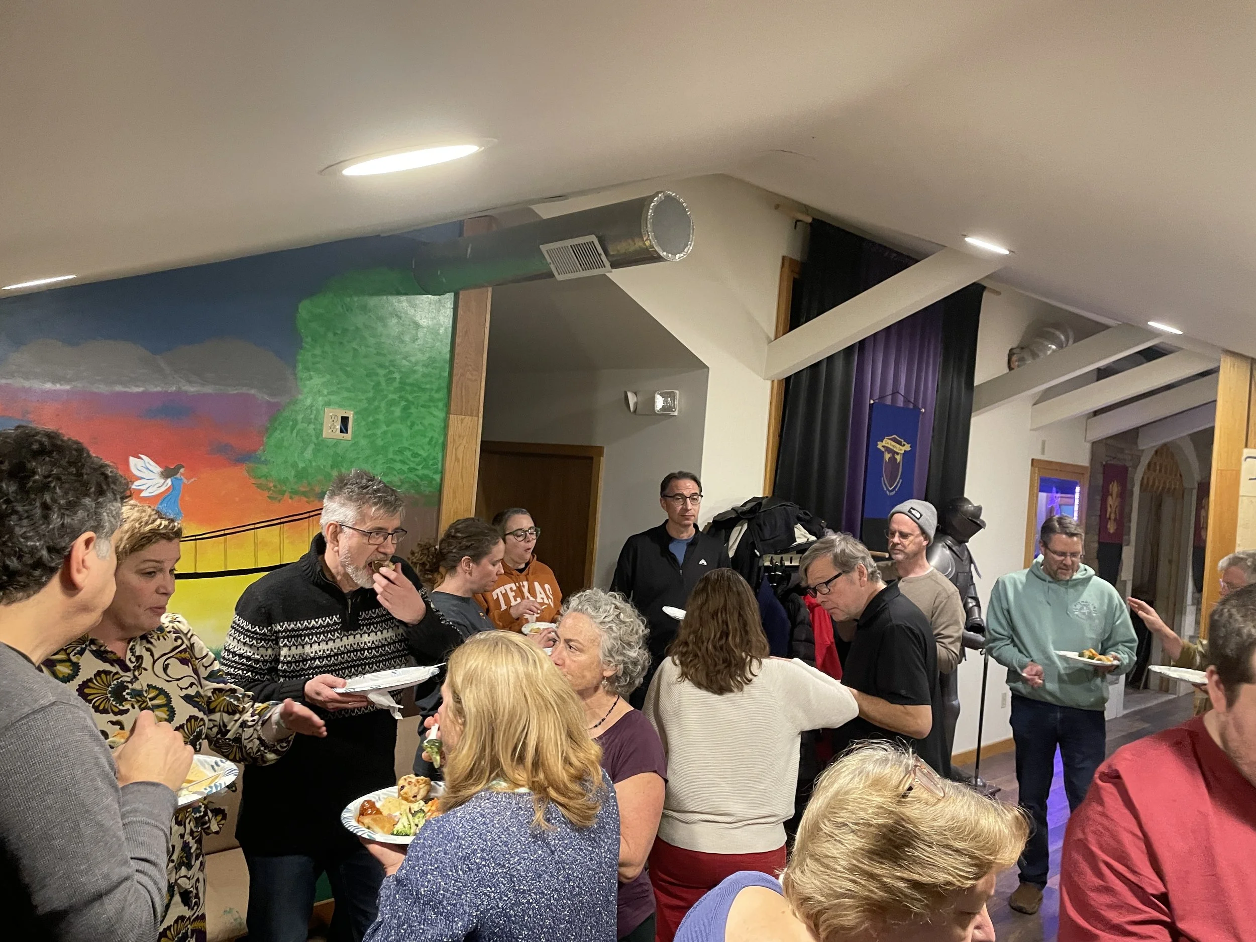 Indoor gathering of people at a social event, some holding plates of food, in a room with colorful mural on the wall and black curtains, with a few individuals wearing casual clothing and glasses.