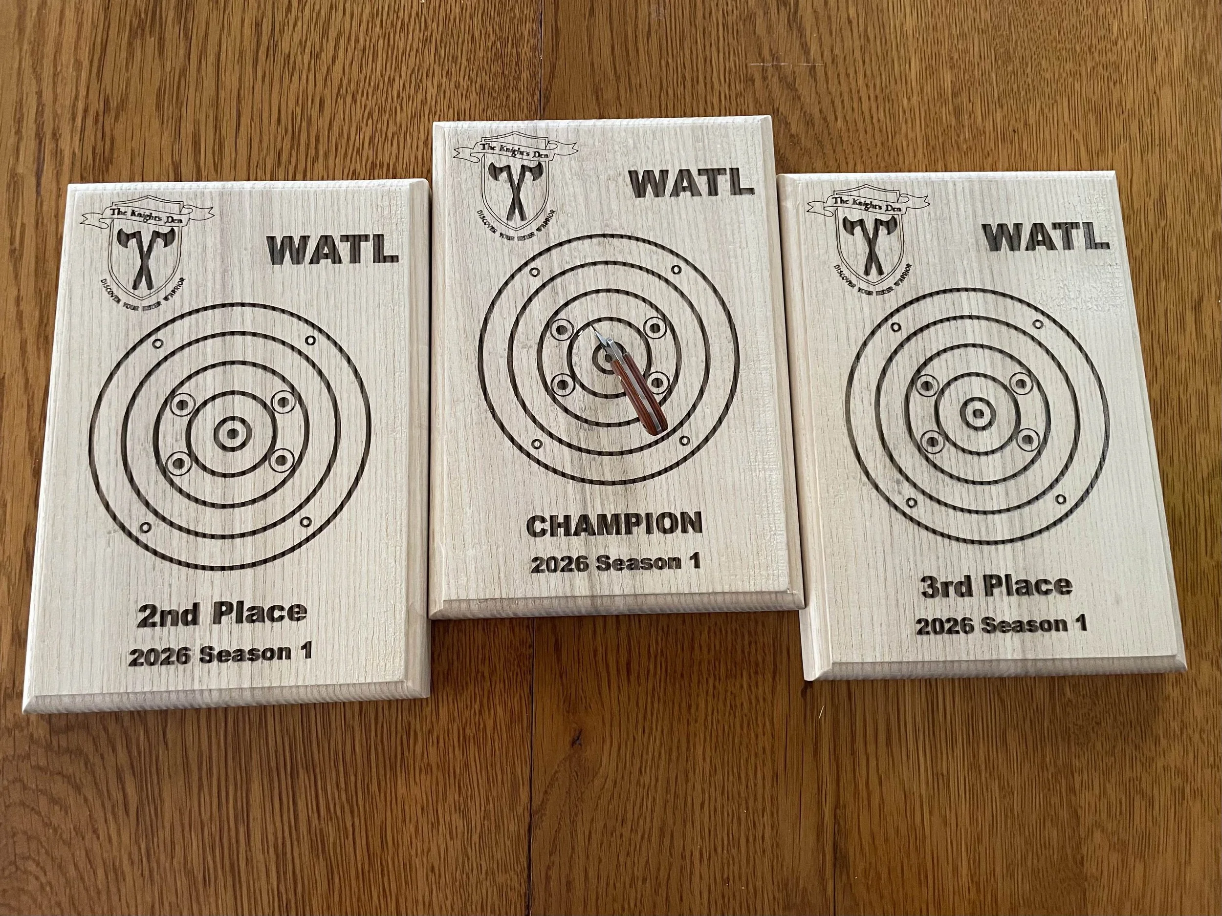 Three wooden axe throwing target plaques, the middle one marked as champion, flanked by second and third place, all from the 2026 season, with the middle plaque having a mini axe.