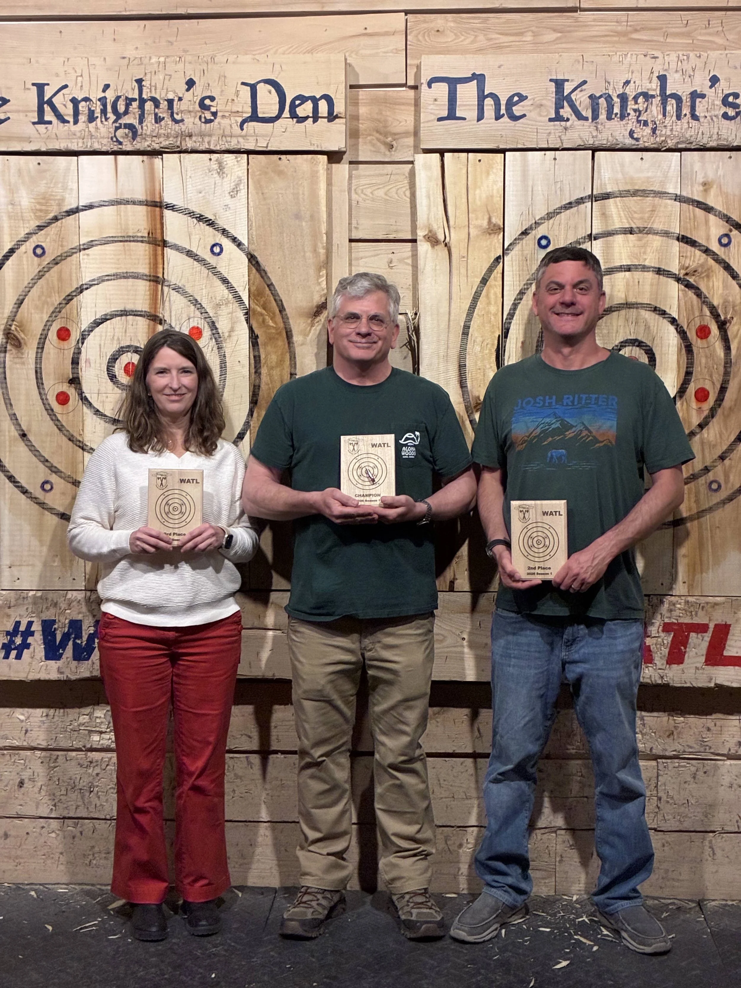 Three people standing in front of a wooden axe throwing target backdrop at the end of season axe throwing tournament. They are holding their plaques for first, second, and third place.