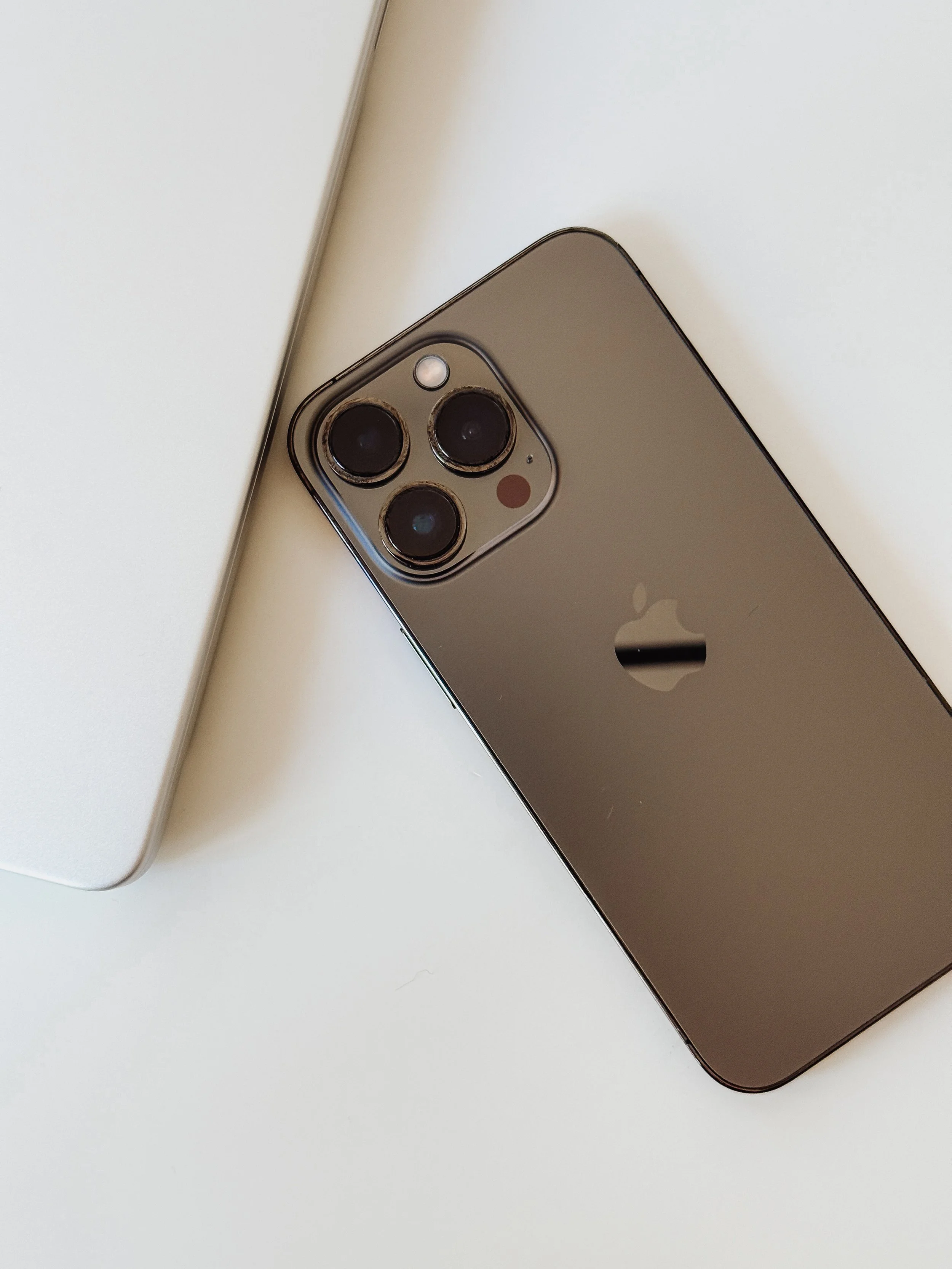 Close-up of a silver iPhone with three camera lenses on the top left corner, placed on a white surface next to a white laptop.