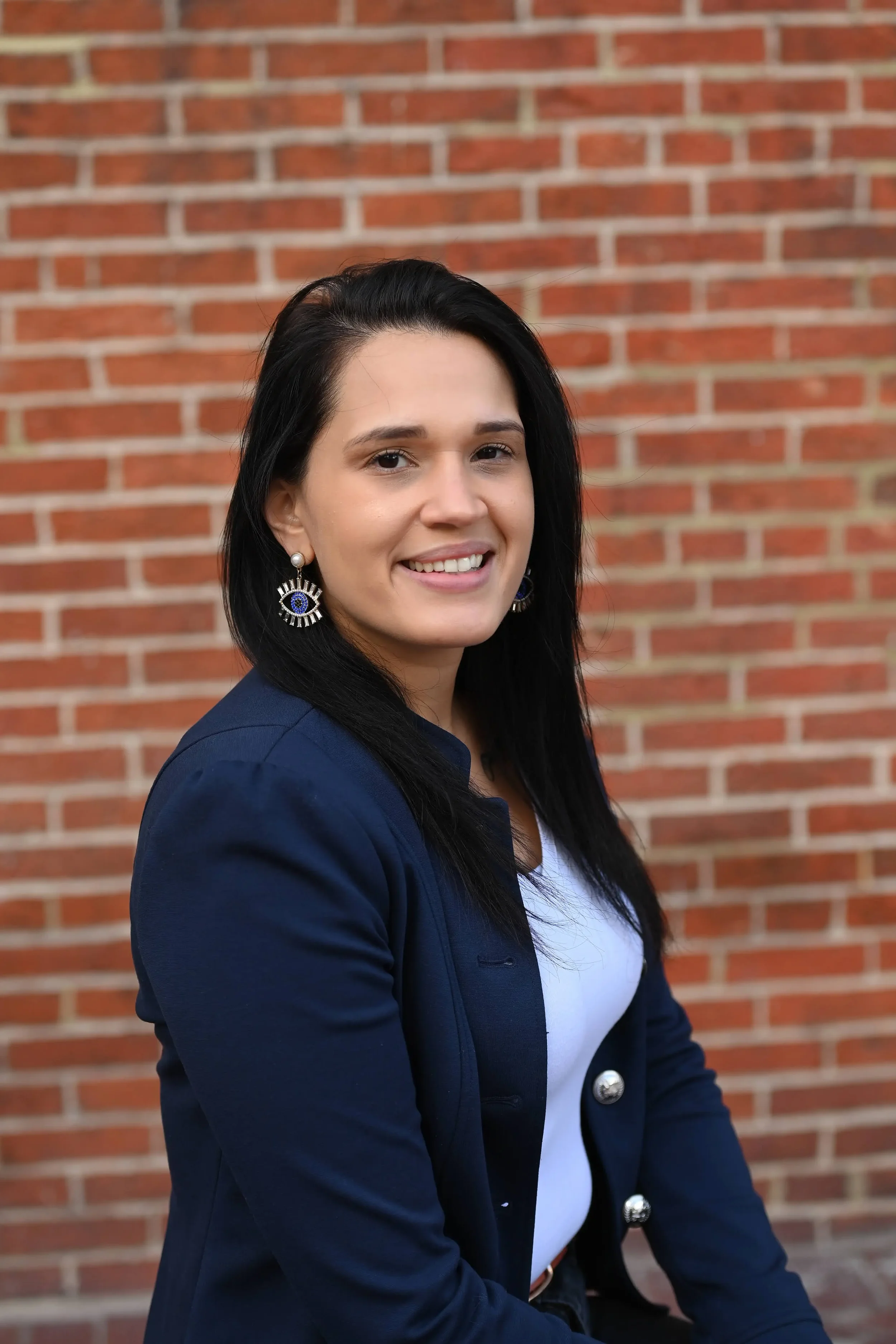A woman with long black hair wearing a navy blazer, white top, and eye-shaped earrings, sitting outdoors in front of a red brick wall.