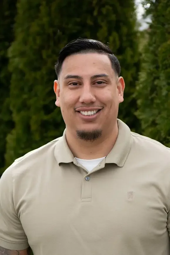 A man smiling outdoors with green trees in the background, wearing a beige polo shirt.