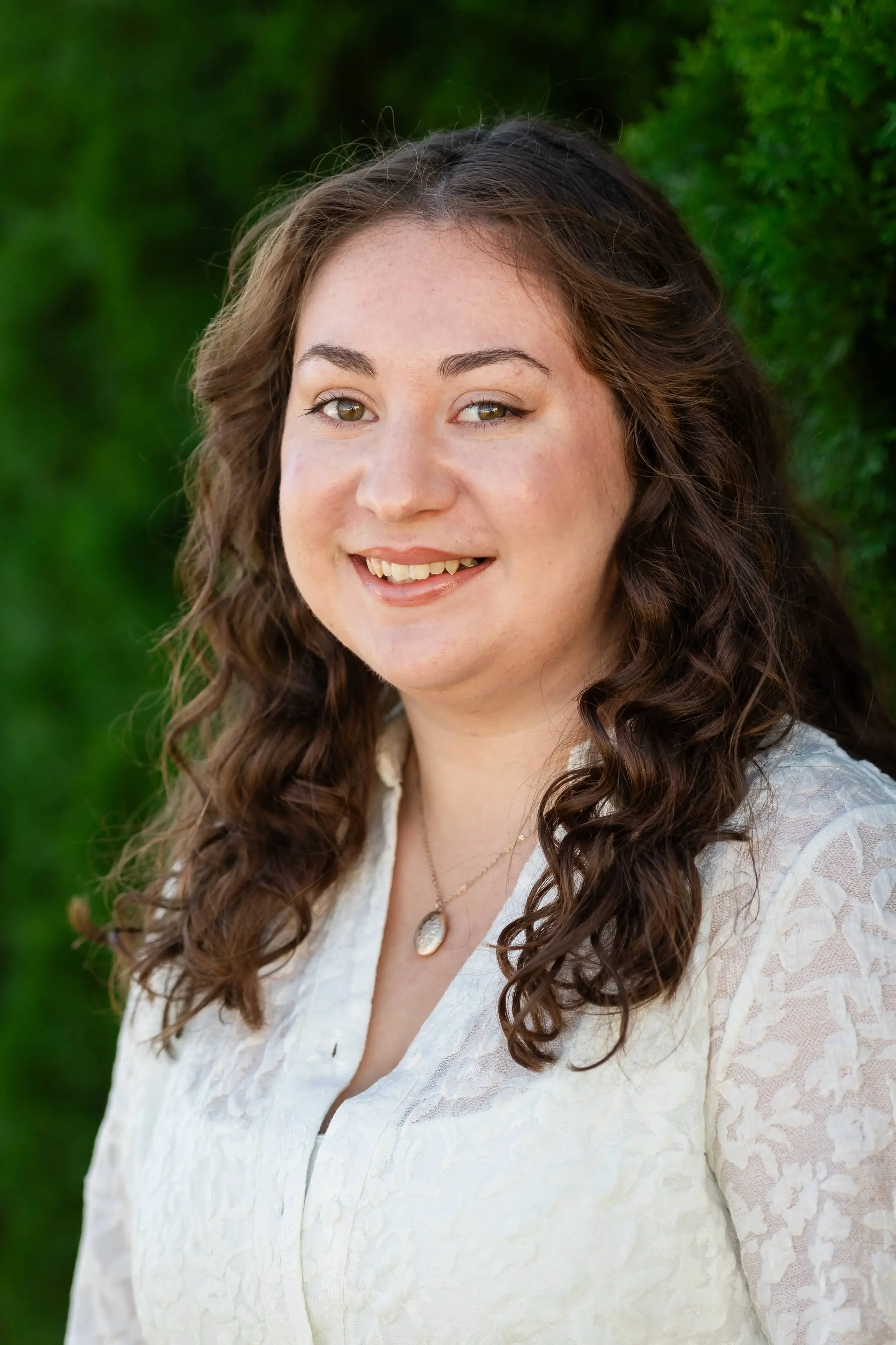 A woman with long curly brown hair and light skin, wearing a white lace blouse and a gold necklace with a pendant, standing outdoors with green foliage in the background.
