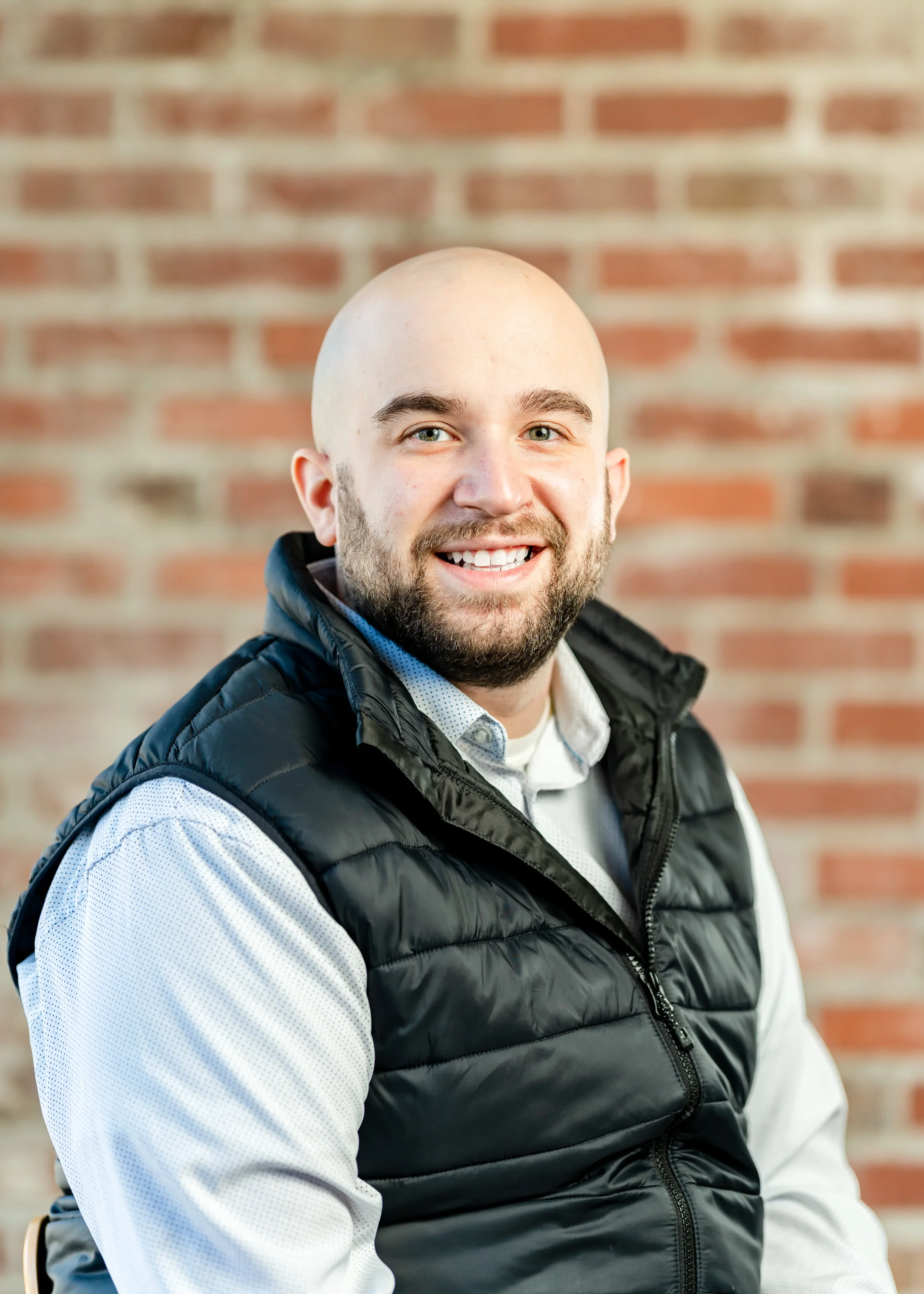 Portrait of a smiling bald man with a beard, wearing a light-colored shirt and black puffer vest, against a brick wall background.