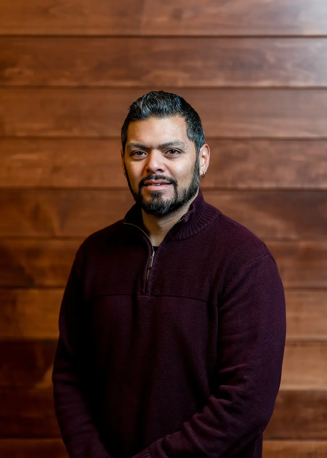 A man with dark hair, beard, and earring, wearing a maroon sweater, standing against a wooden background.